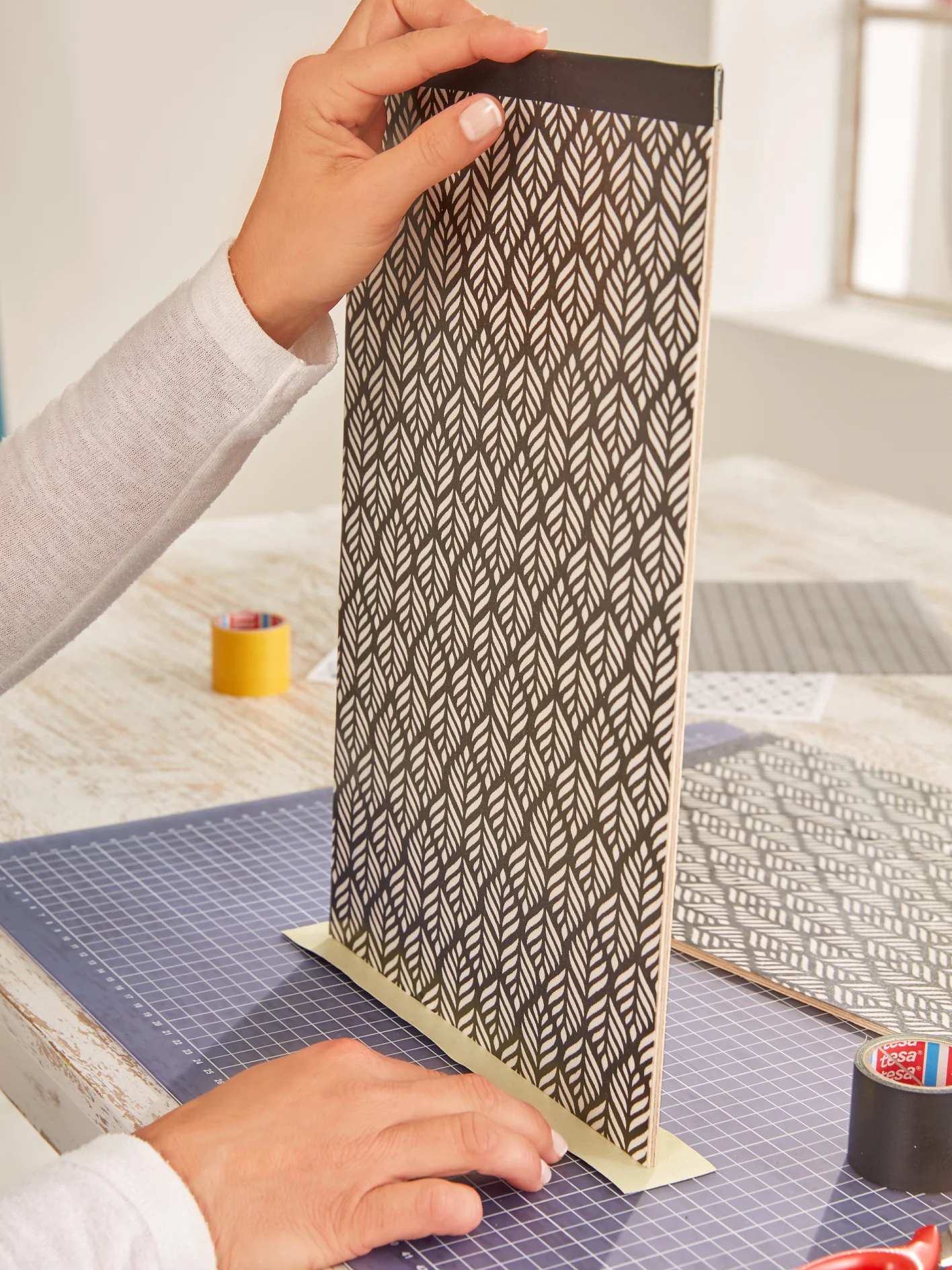 A person is holding a vertically positioned black and white patterned board on a blue cutting mat. Various crafting materials, including tesa tape and fabric, are on a wooden table. (This text has been generated by AI)