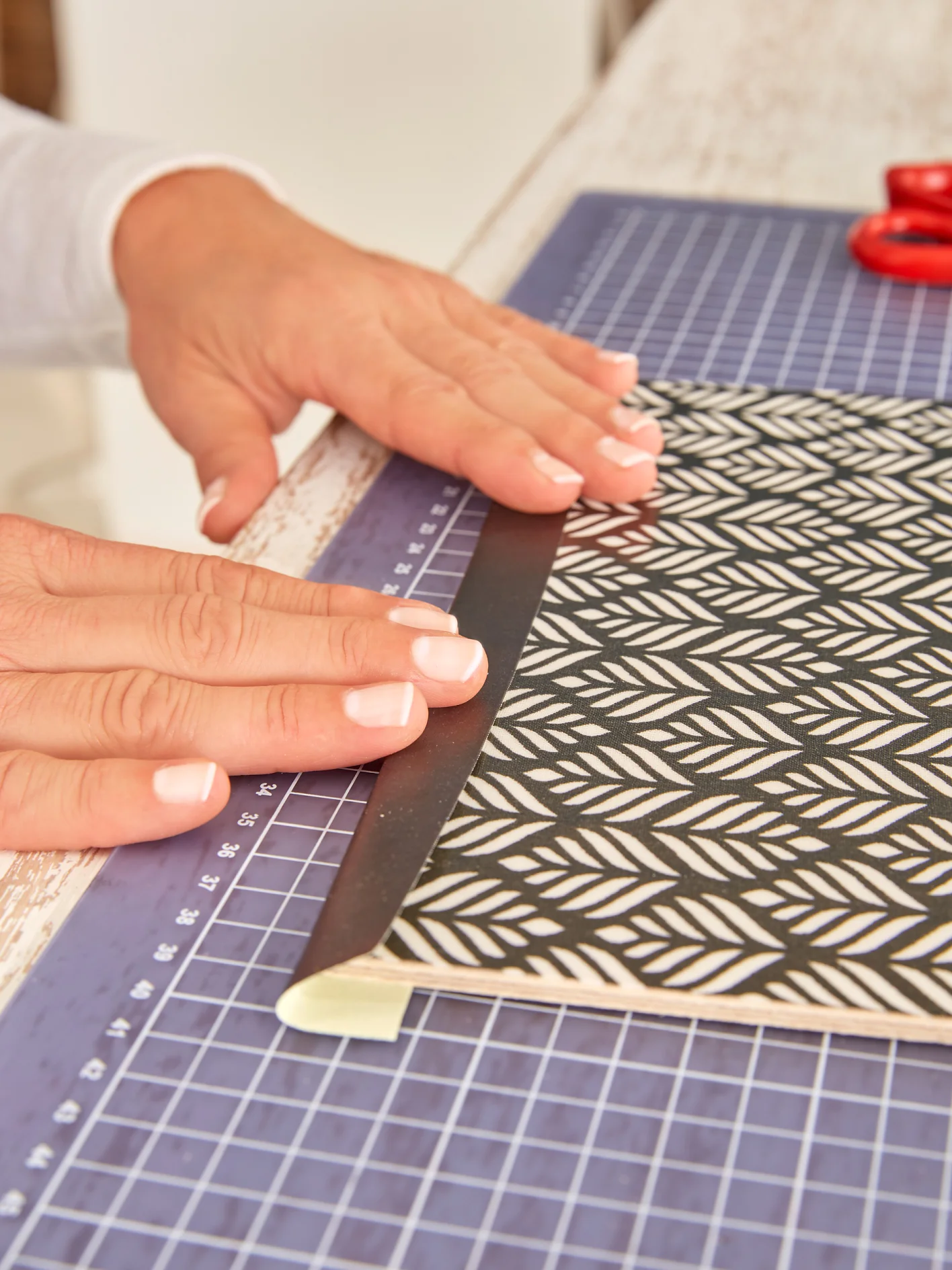 Hands are folding the cover of a handmade notebook with a black and white leaf pattern. The notebook is on a cutting mat with a grid design, and a pair of red scissors is visible in the upper right corner, along with tesa tape positioned nearby for securing materials. (This text has been generated by AI)
