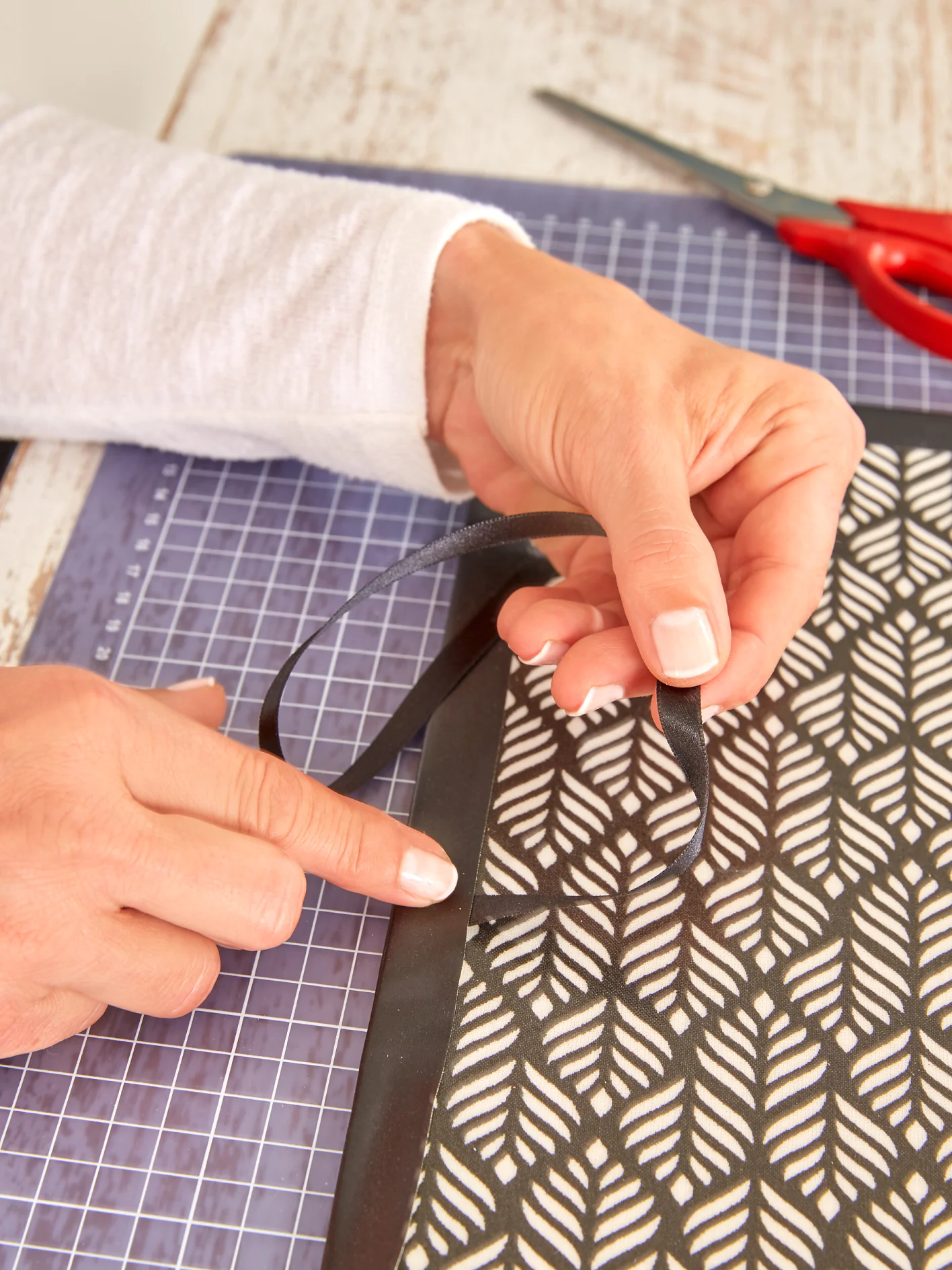 A person is working on a craft project. They are attaching a black ribbon to the edge of a decorative sheet with a leaf pattern using tesa tape. There is a cutting mat underneath and red scissors nearby on the wooden table. (This text has been generated by AI)