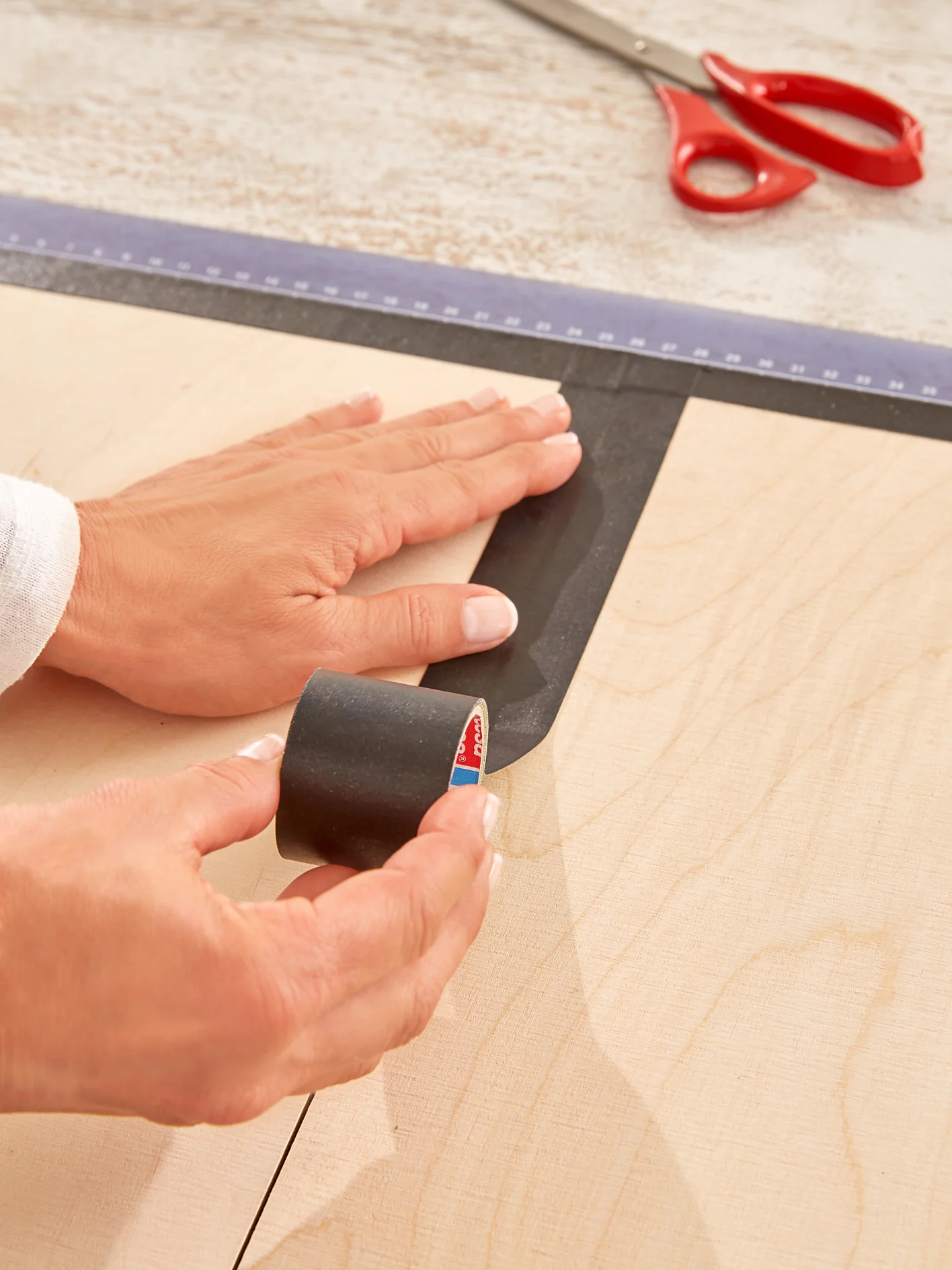 A person applies black tesa tape onto a wooden surface using their hand and a roll of tesa tape. A ruler and a pair of red scissors are visible in the background on a light-colored table. (This text has been generated by AI)
