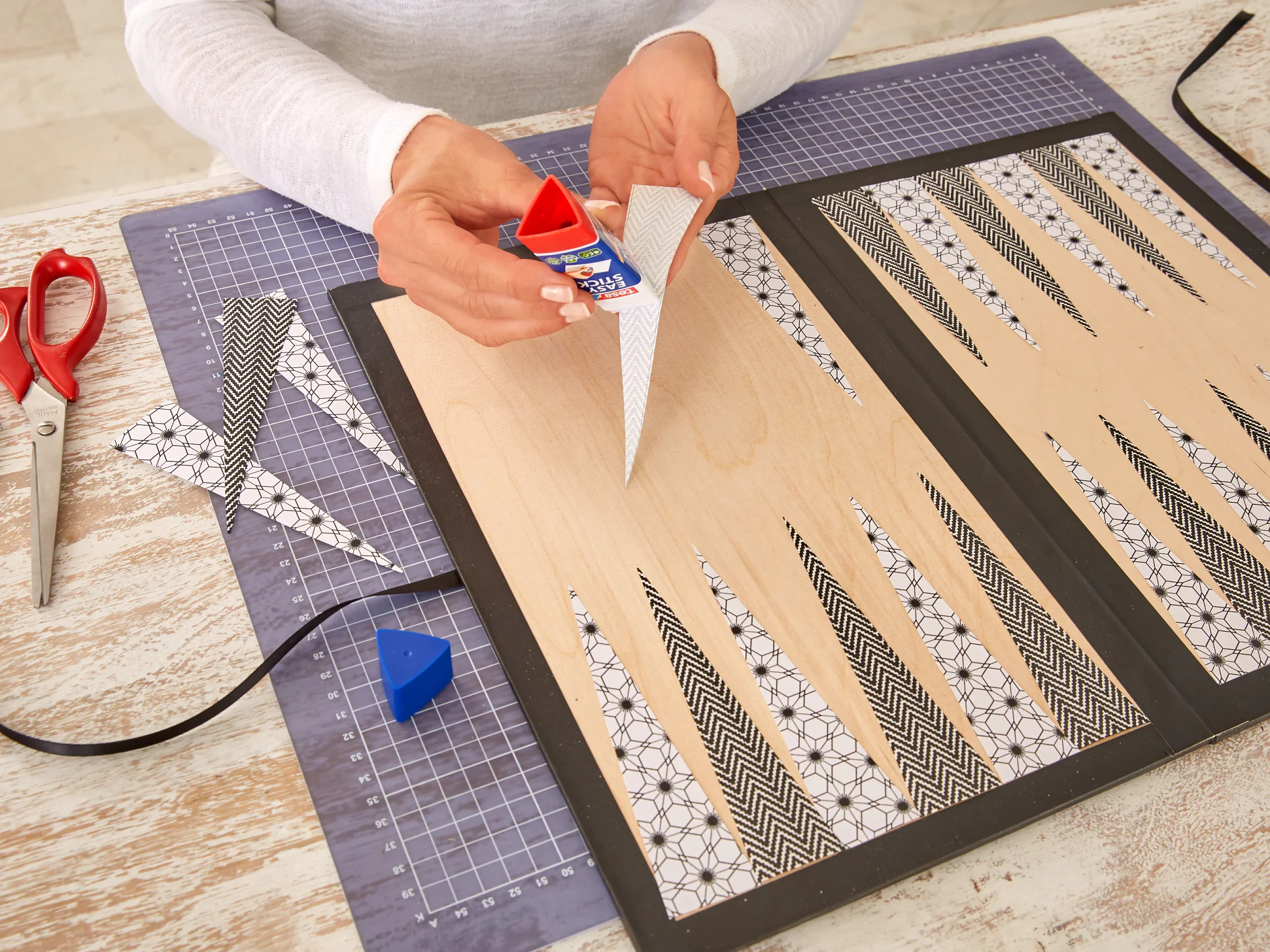 A person is applying glue to a triangular piece of paper with a tesa tape glue stick. The paper features black and white patterns. The workspace includes a cutting mat, scissors, and other patterned paper pieces arranged on a wooden surface. (This text has been generated by AI)
