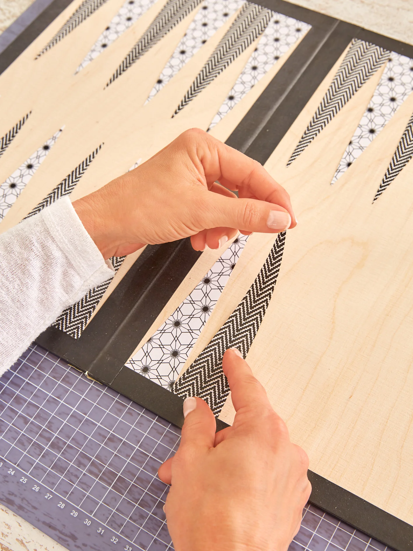 Hands are placing patterned strips of tesa tape onto a wooden surface. The background includes a grid cutting mat, and the strips have various black and white geometric designs. (This text has been generated by AI)