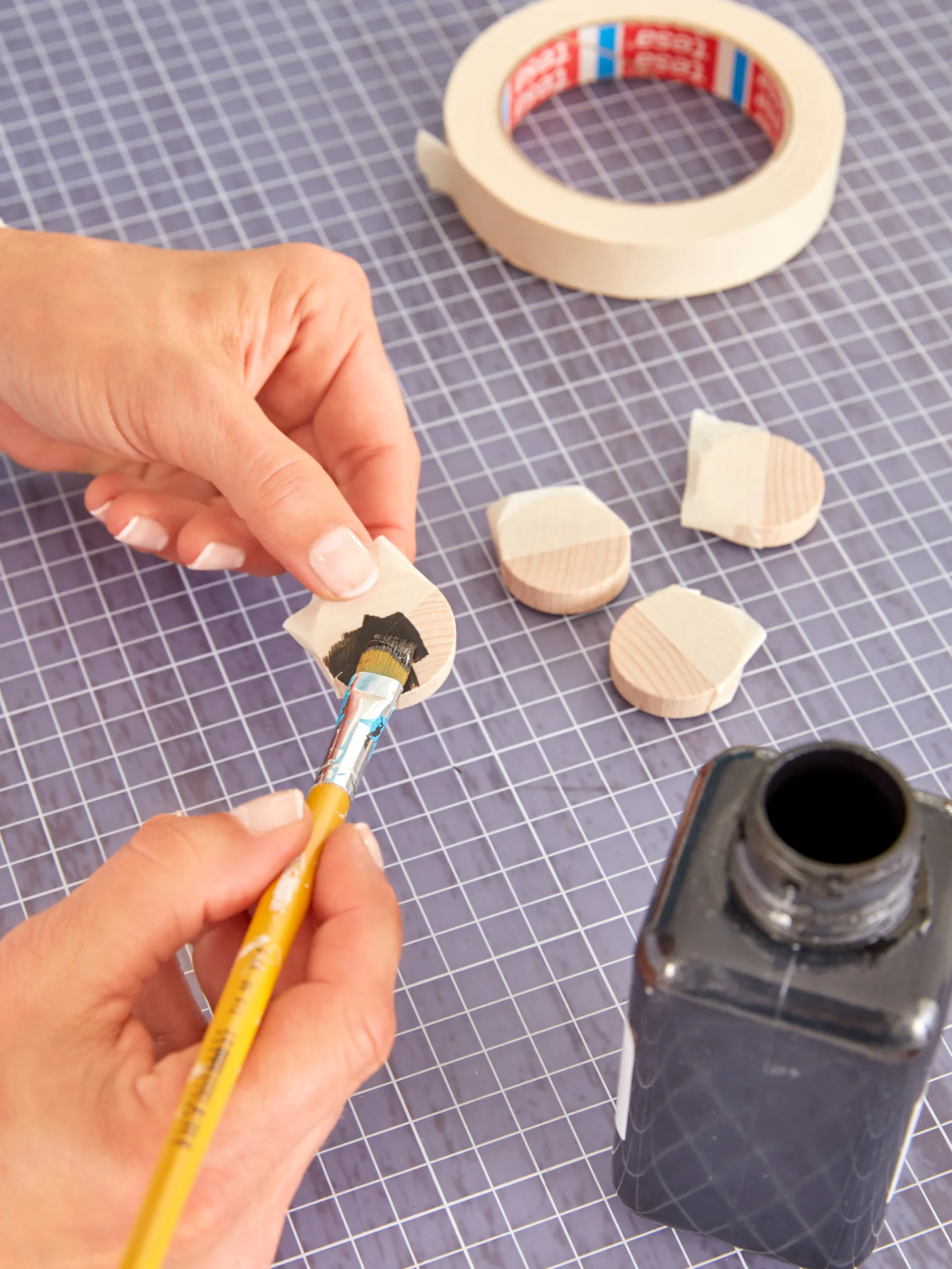 A person is painting small wooden circles with black paint using a brush. The circles are placed on a grid-patterned surface. A roll of tesa tape and an open black paint container are visible in the background. (This text has been generated by AI)