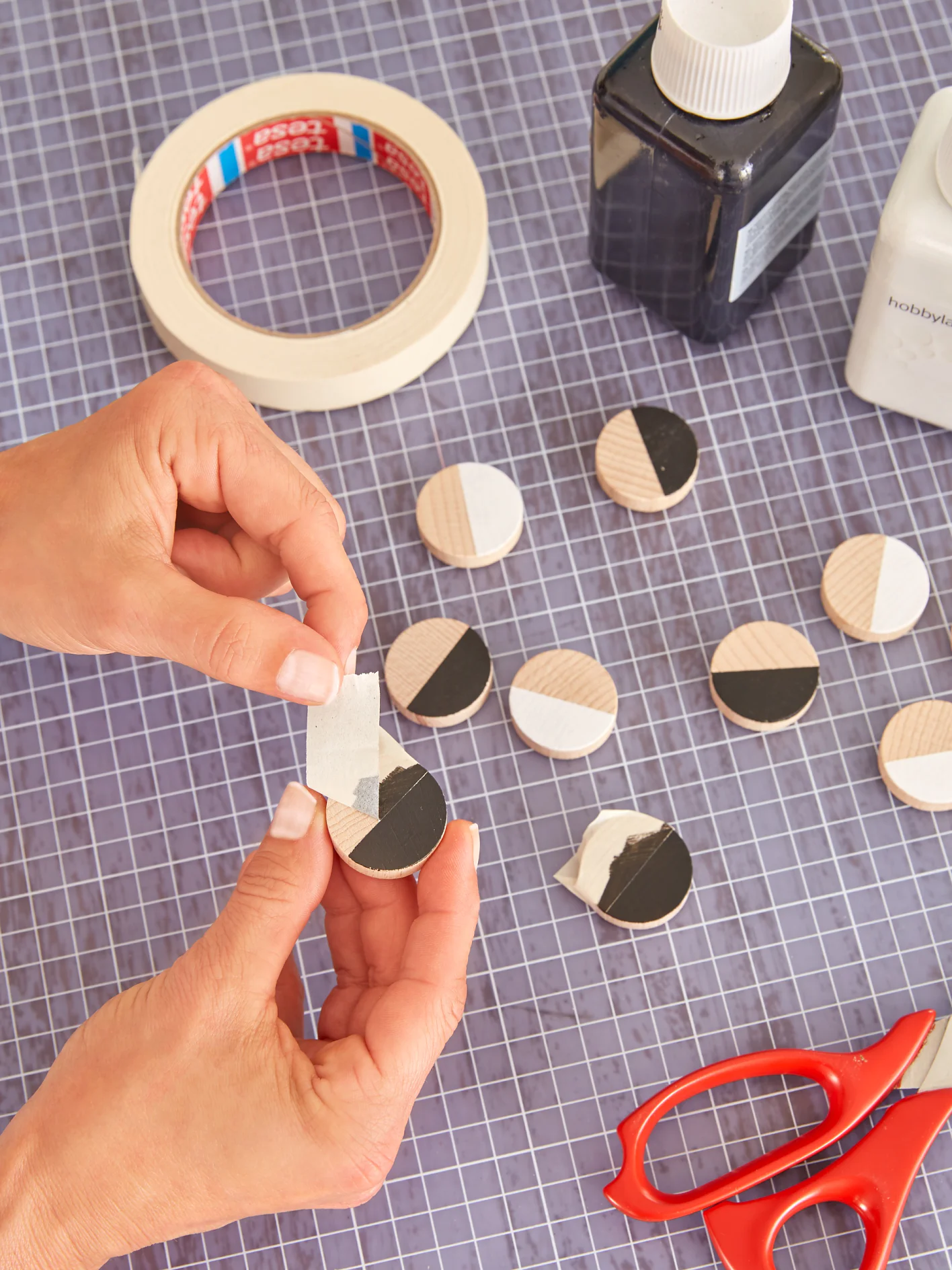 Hands are seen applying tesa tape to small, round wooden pieces painted in black and white halves. A roll of tesa tape, a black bottle, a white bottle, and red scissors are on a grid-patterned surface. (This text has been generated by AI)