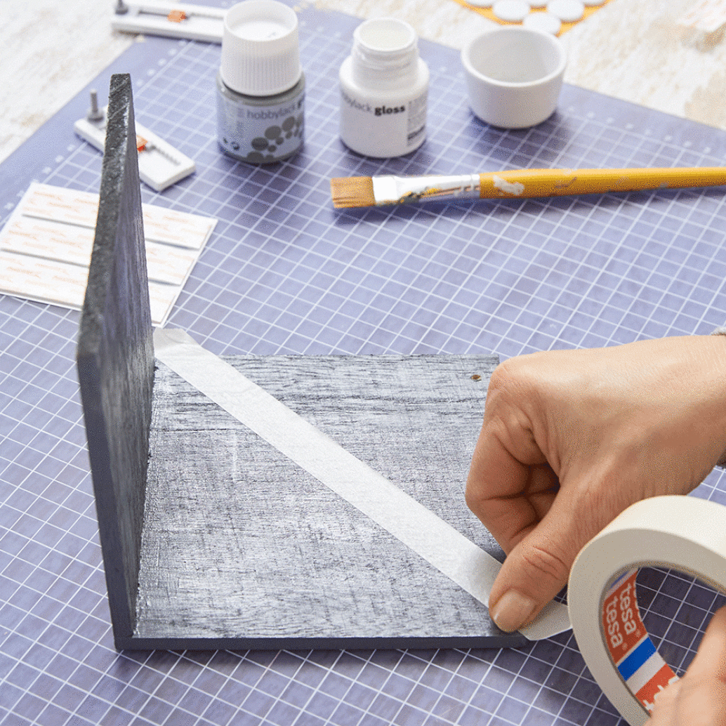 A person applies tesa tape to a painted wooden L-shaped bracket on a crafting table. Nearby are small bottles of paint, a paintbrush, and various crafting supplies. The table surface features a blue cutting mat. (This text has been generated by AI)