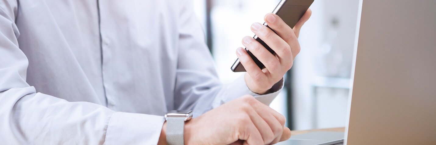 白いシャツを着た人物が机に座り、右手にスマートフォンを持ち、左手には開いたノートパソコンを置いています。背景は窓から差し込む自然光で柔らかく照らされています。. (This text has been generated by AI)