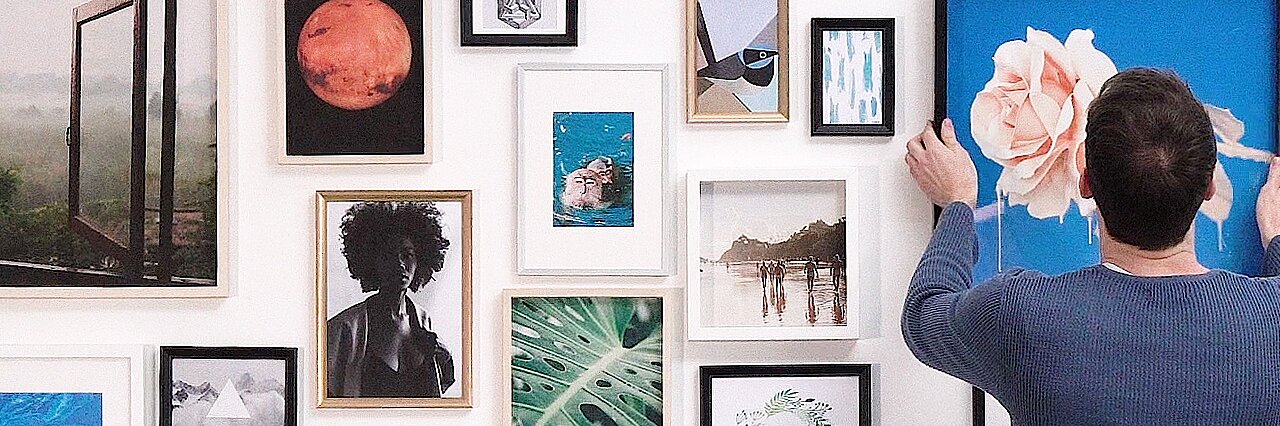 A person in a blue sweater is adjusting a framed picture of a large rose on a blue background on a gallery wall using tesa tape. The wall features various framed artworks, including a red planet, landscapes, abstract designs, and nature images. (This text has been generated by AI)