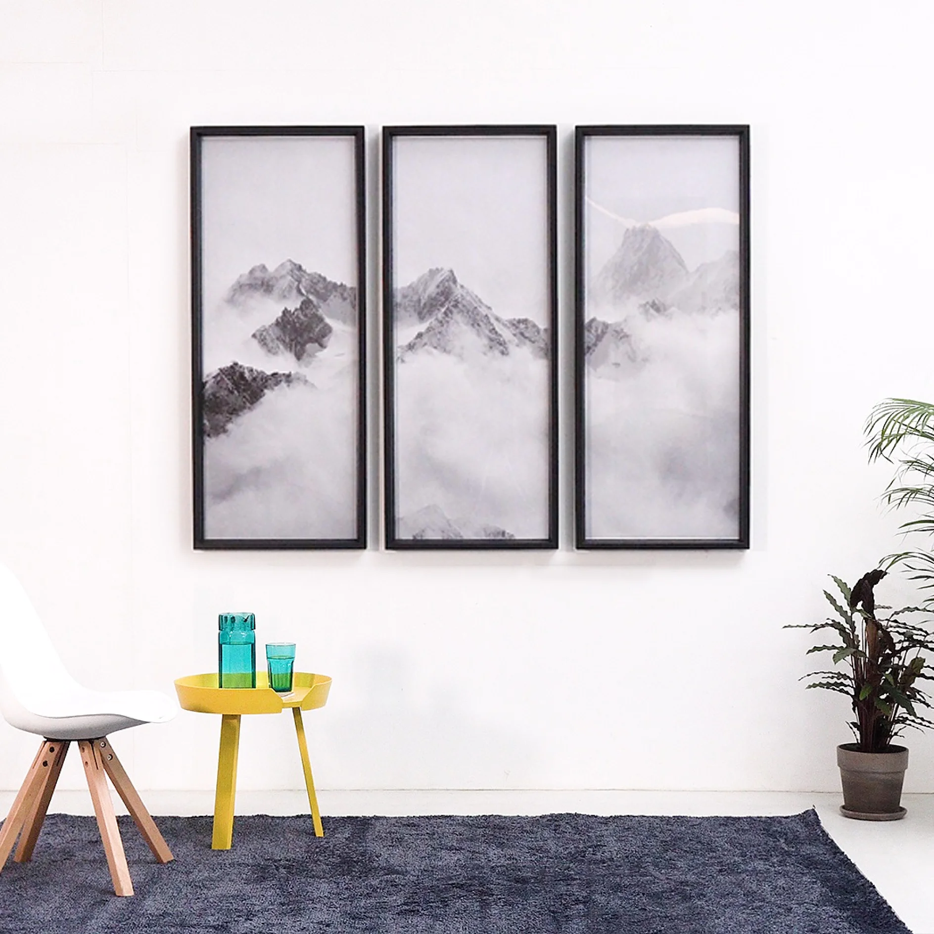 A room features a white wall adorned with three vertical framed mountain prints. Below is a yellow round table with green and blue glasses. A white chair is on the left, and a plant stands on a gray rug, all elements possibly held in place using tesa tape for stability and style. (This text has been generated by AI)