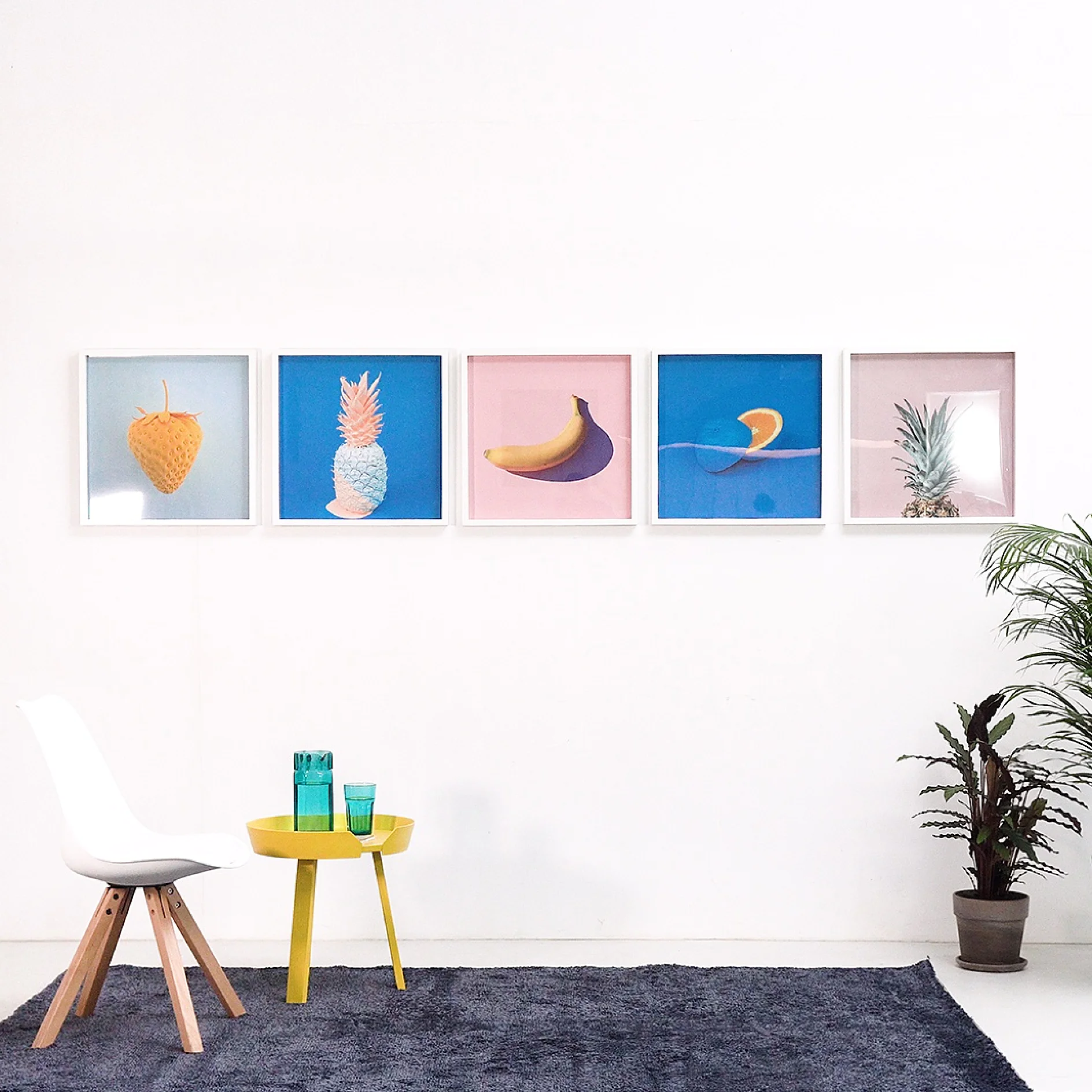 A minimalist room features a series of five framed fruit prints on a white wall, all secured using tesa tape. Below, theres a white chair, a small yellow table with two teal glasses, and a green plant. The floor is covered by a dark rug. (This text has been generated by AI)