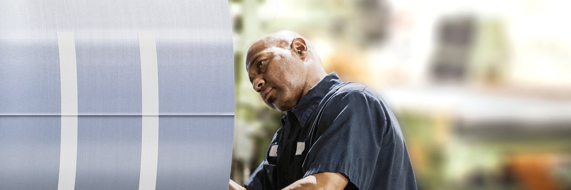 A worker in a blue uniform is inspecting or working on a large roll of tesa tape in an industrial setting. The background is blurred, highlighting the focus on the worker and the roll of tesa tape.