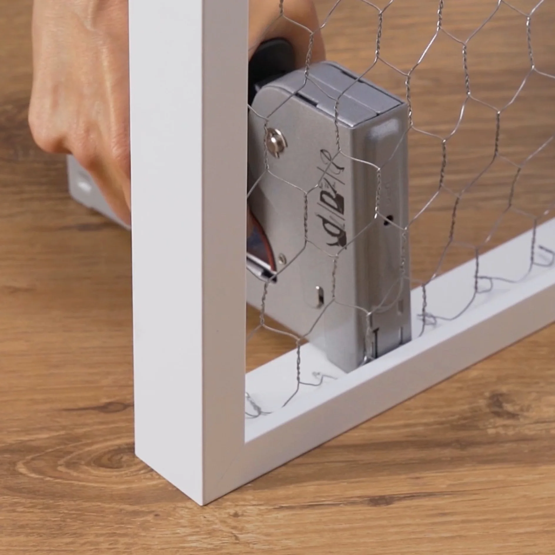 A hand using a staple gun to attach a piece of chicken wire to a white wooden frame, set on a wooden surface with tesa tape. (This text has been generated by AI)