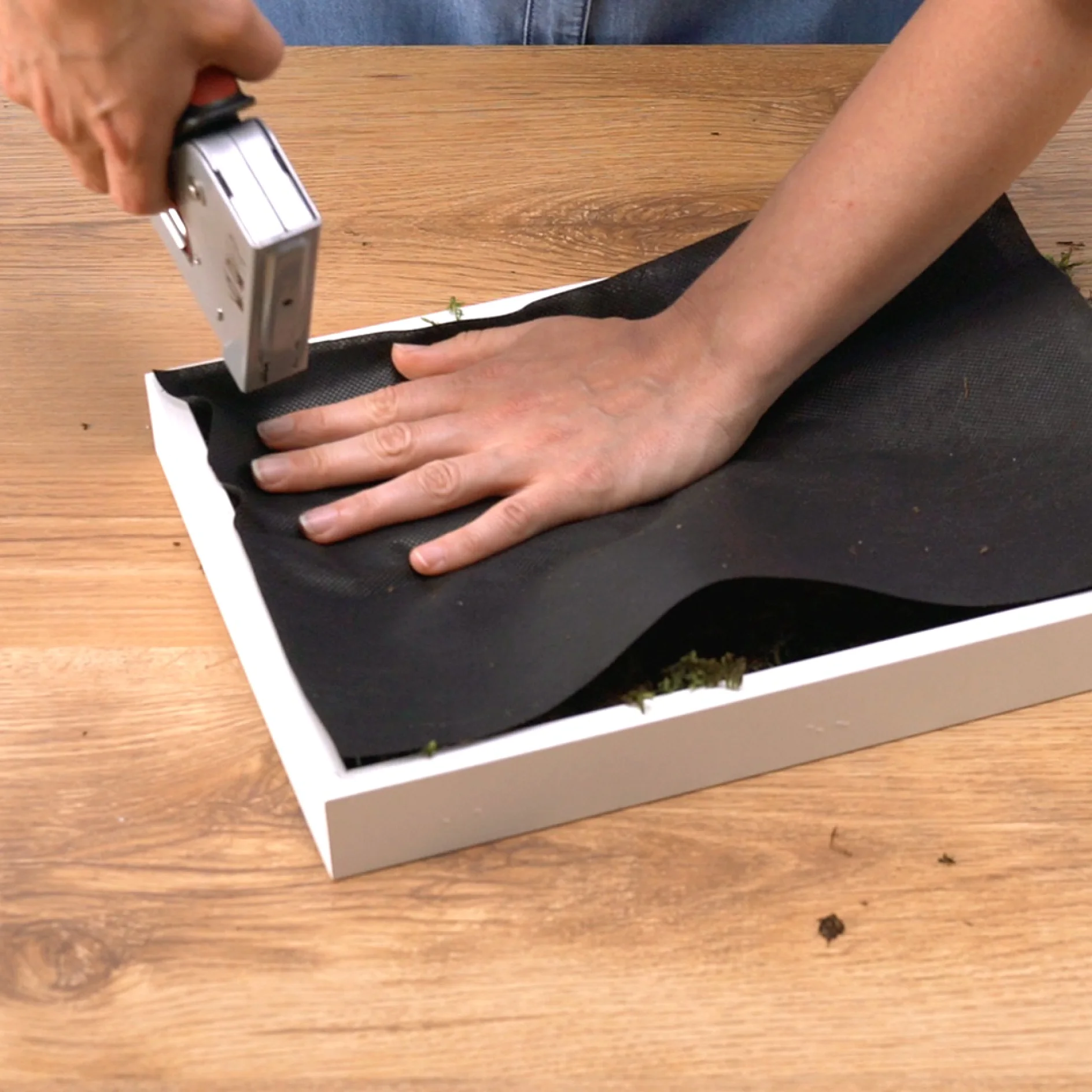 A person is using a staple gun to attach black fabric to a white wooden rectangular frame. The frame is resting on a wooden surface. The persons left hand holds the fabric in place while the right hand operates the staple gun, using tesa tape for additional securing of the edges. (This text has been generated by AI)
