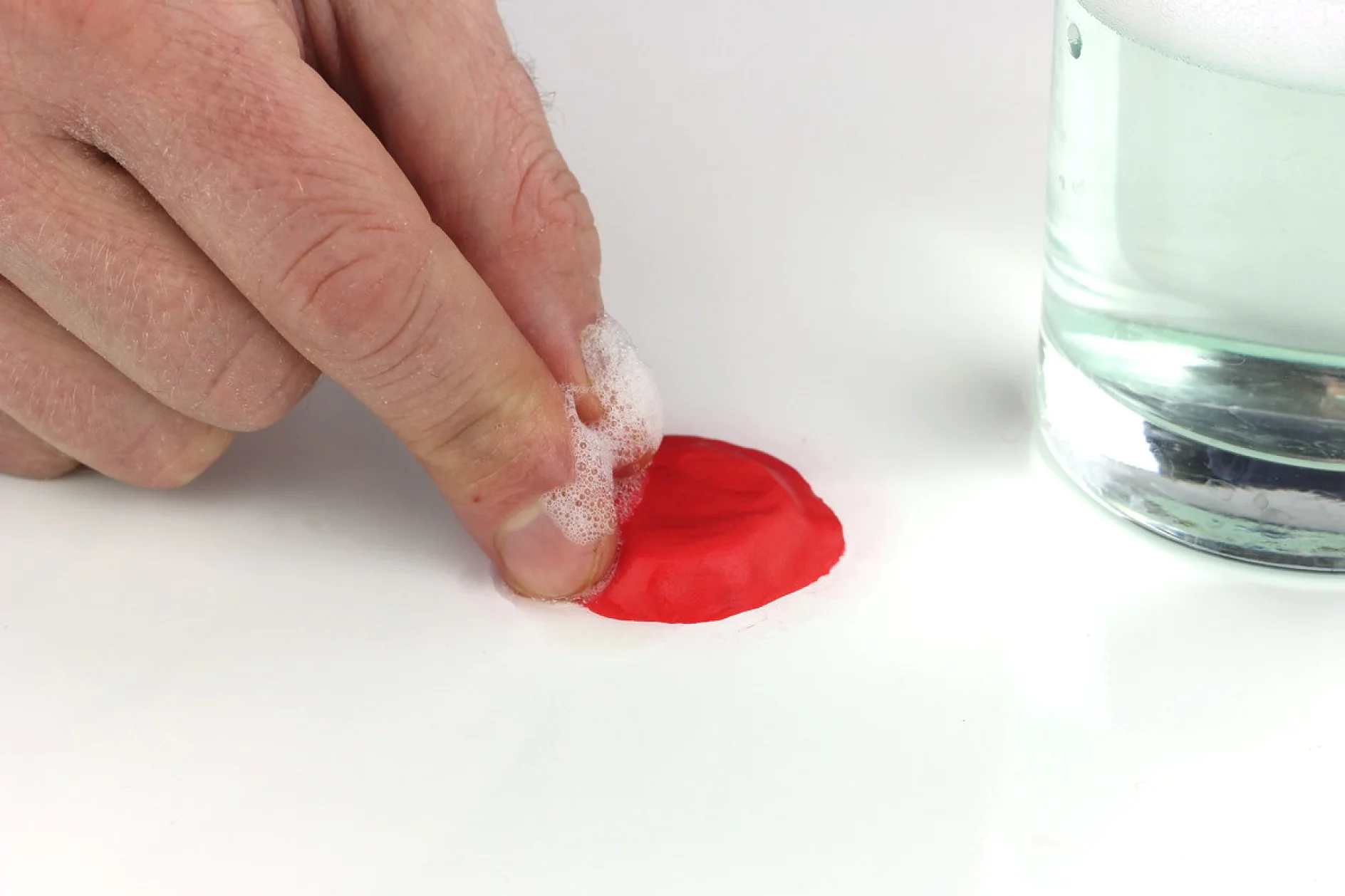 手は石鹸水を含ませたスポンジを使って、テサテープを貼った白い表面の赤い斑点を拭いています。透明な液体が入ったグラスが近くにあります。. (This text has been generated by AI)