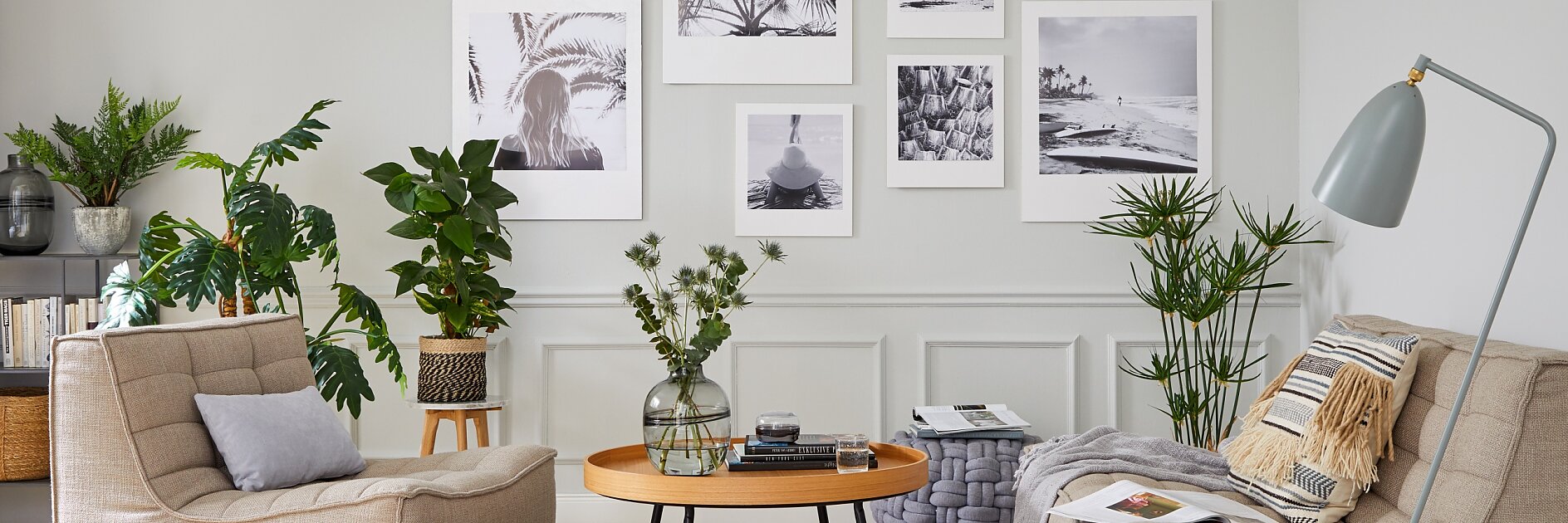 A cozy living room with beige armchairs and a round wooden coffee table. The walls display black and white photos in white frames, all secured with tesa tape. Potted plants are scattered around, and a grey floor lamp stands to the right. A woven basket and side table are also present. (This text has been generated by AI)