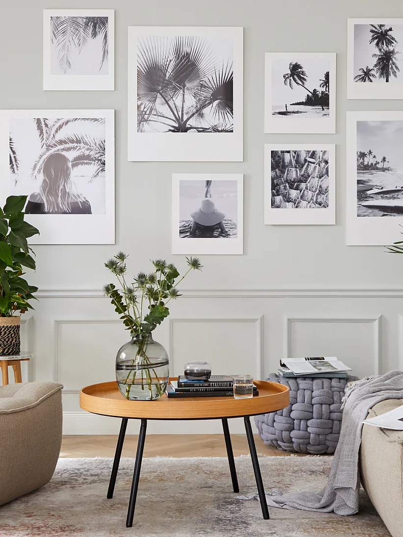 A cozy living room with beige armchairs and a round wooden coffee table. The walls display black and white photos in white frames, all secured with tesa tape. Potted plants are scattered around, and a grey floor lamp stands to the right. A woven basket and side table are also present. (This text has been generated by AI)
