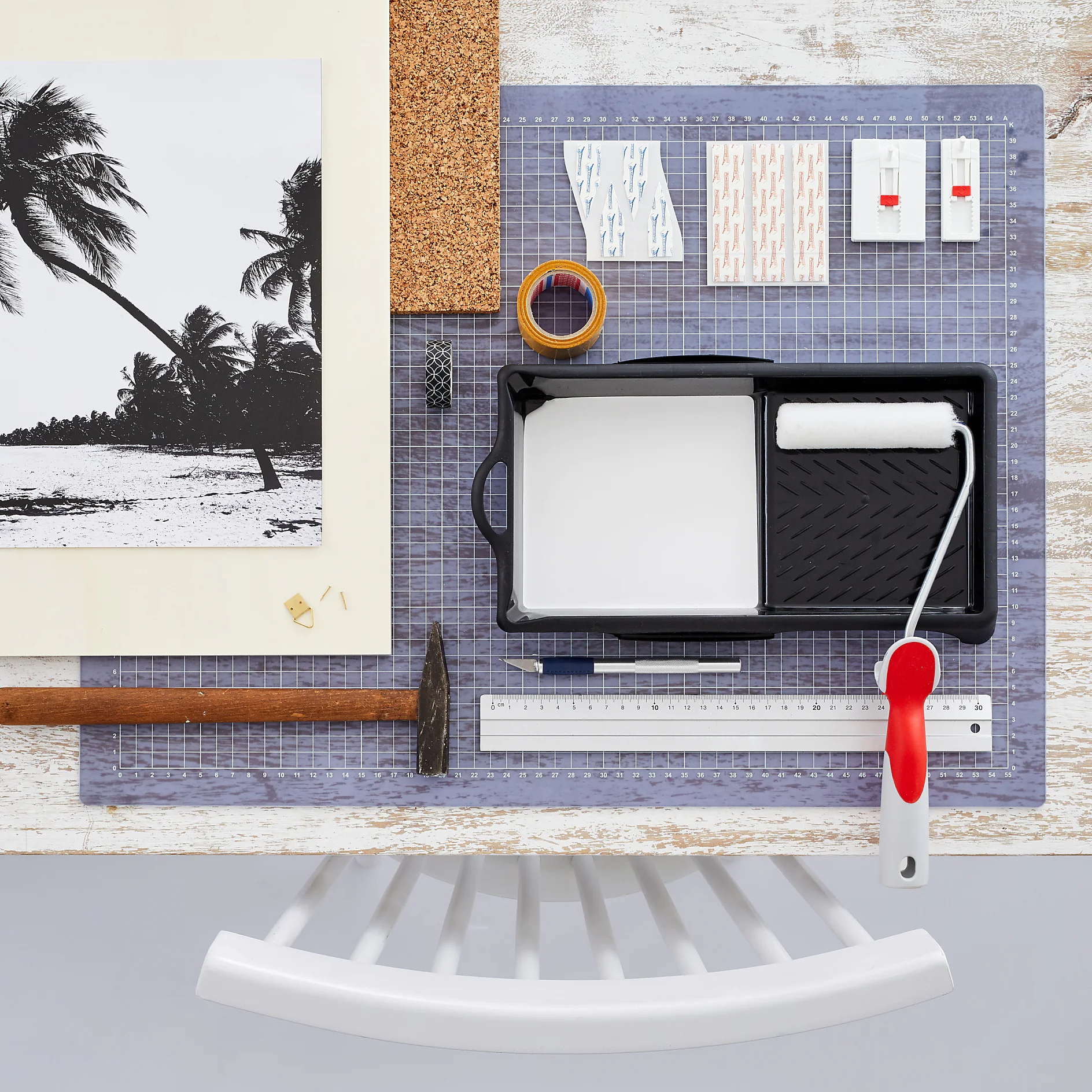 A paint roller set with a red handle lies in a tray on a desk, alongside a hammer, measuring tesa tape, ruler, adhesive hooks, a postcard of palm trees, and a piece of cork. A white chair is partially visible at the bottom. (This text has been generated by AI)