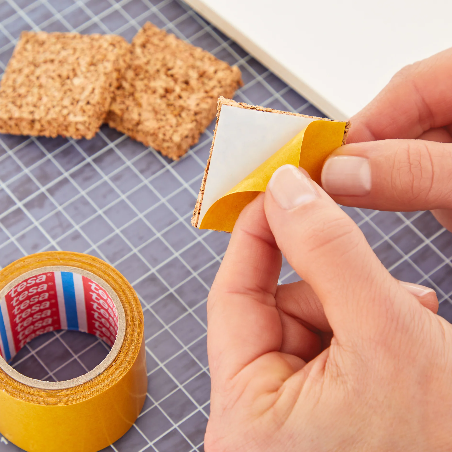 A person is peeling the backing off a small adhesive pad. A roll of tesa tape and additional adhesive pads are on a grid-patterned surface. (This text has been generated by AI)