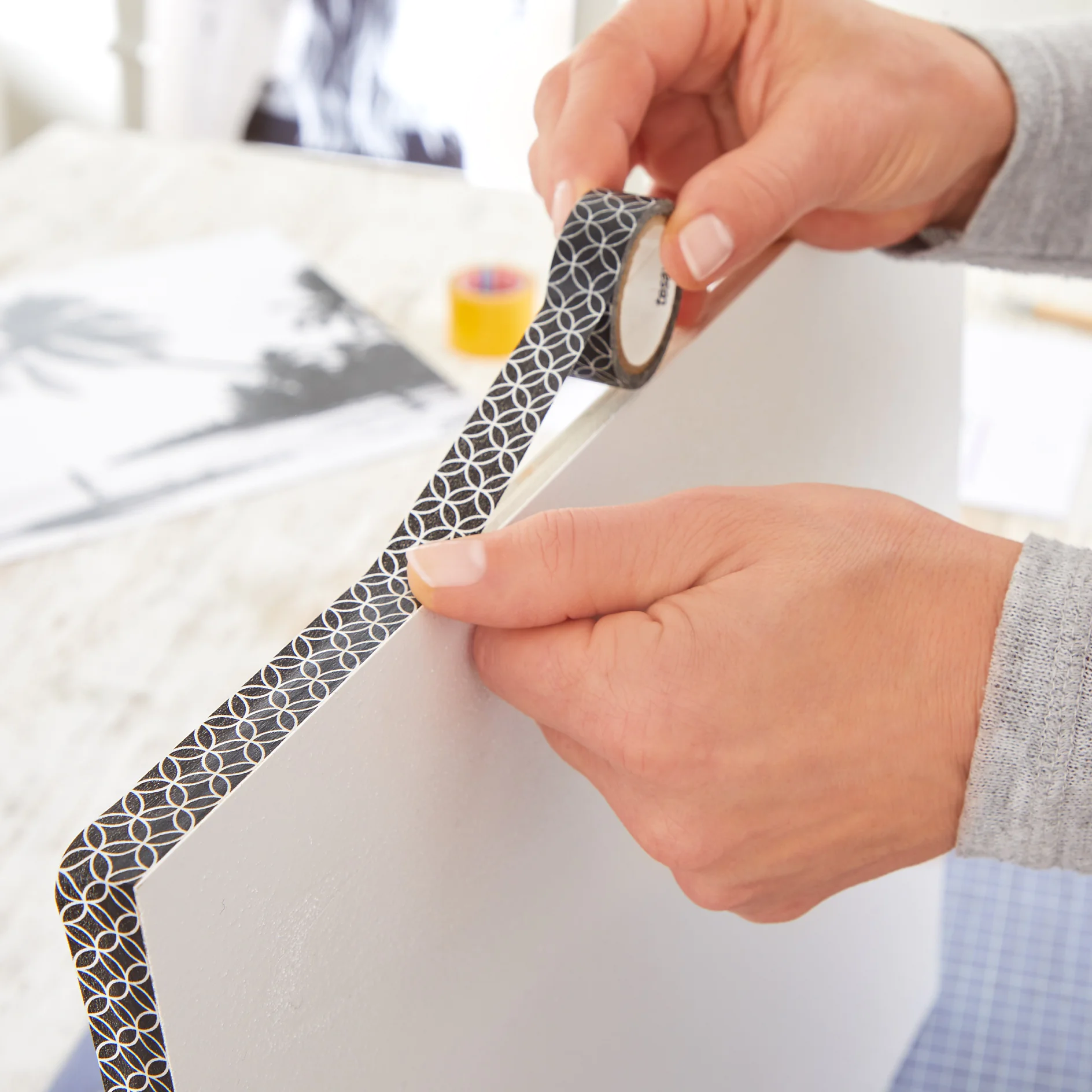 Hands applying patterned black tesa tape to the edge of a white board. A blurred background shows a tabletop with various crafting supplies and a partially visible black-and-white photo. (This text has been generated by AI)
