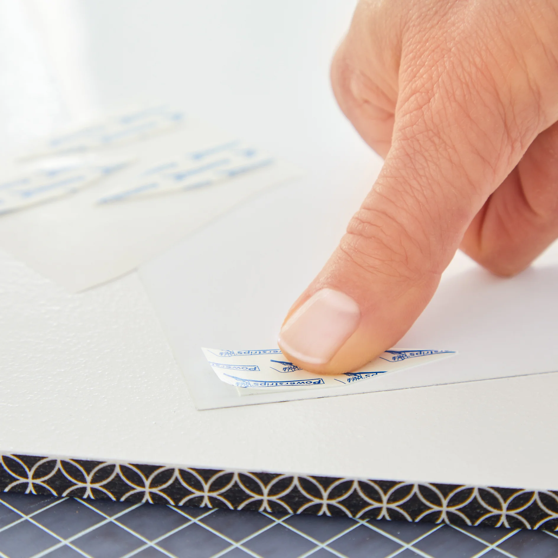 A hand peeling the backing off a small tesa tape strip on a white surface. Several unused tesa tape strips are visible in the background on a sheet. The edge of a decorative item with circular patterns is partially shown below. (This text has been generated by AI)