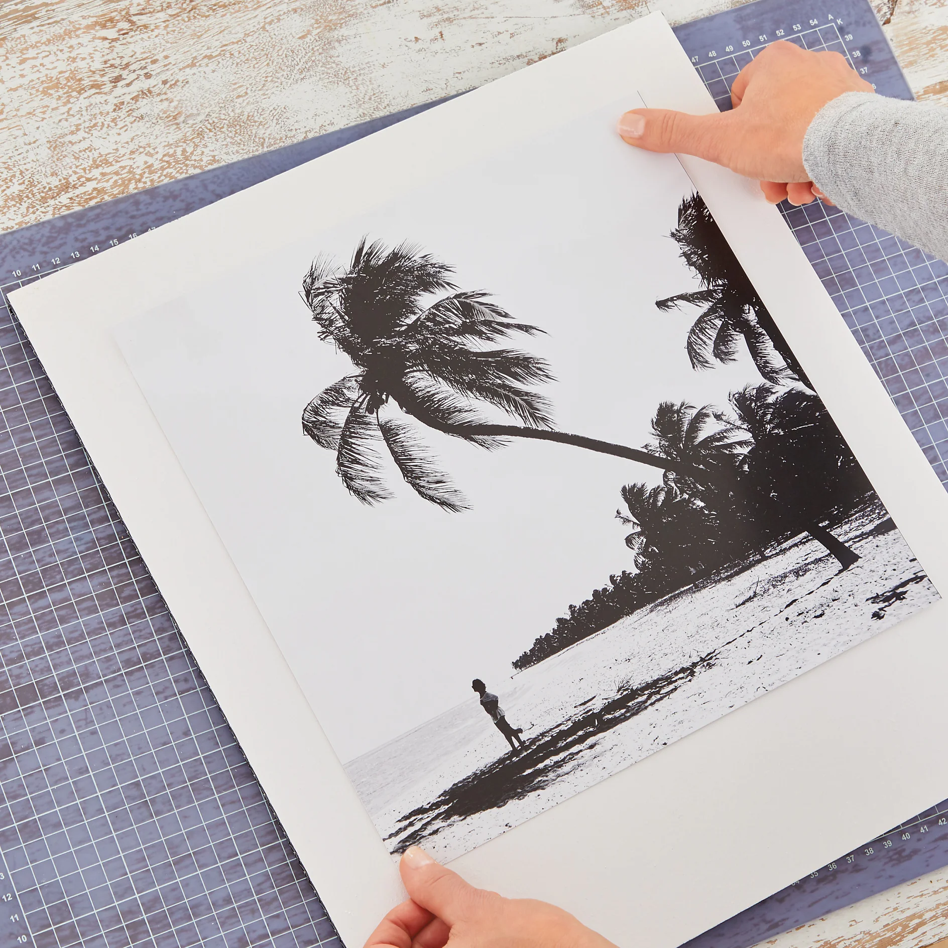 Hands are placing a black-and-white beach photograph featuring palm trees and a person standing by the shore onto a white mat board using tesa tape. The surface beneath is a patterned table. (This text has been generated by AI)