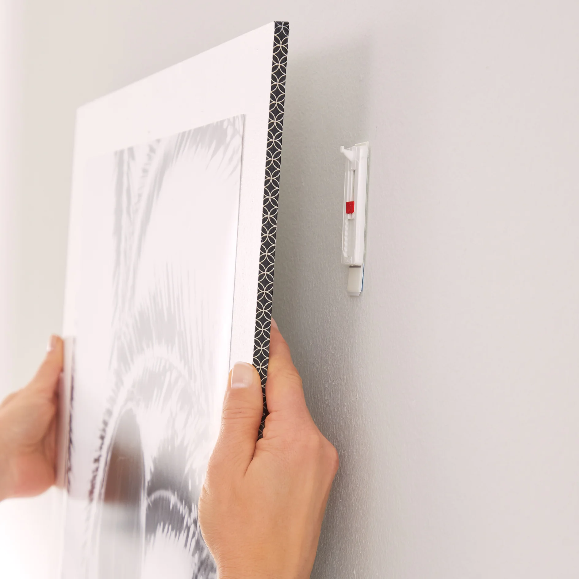 Hands holding a framed picture ready to be hung on a wall. The wall features a hanging hook with a small built-in level indicating the correct orientation. The picture displays a black and white pattern with a thick border, secured using tesa tape for precision and durability. (This text has been generated by AI)