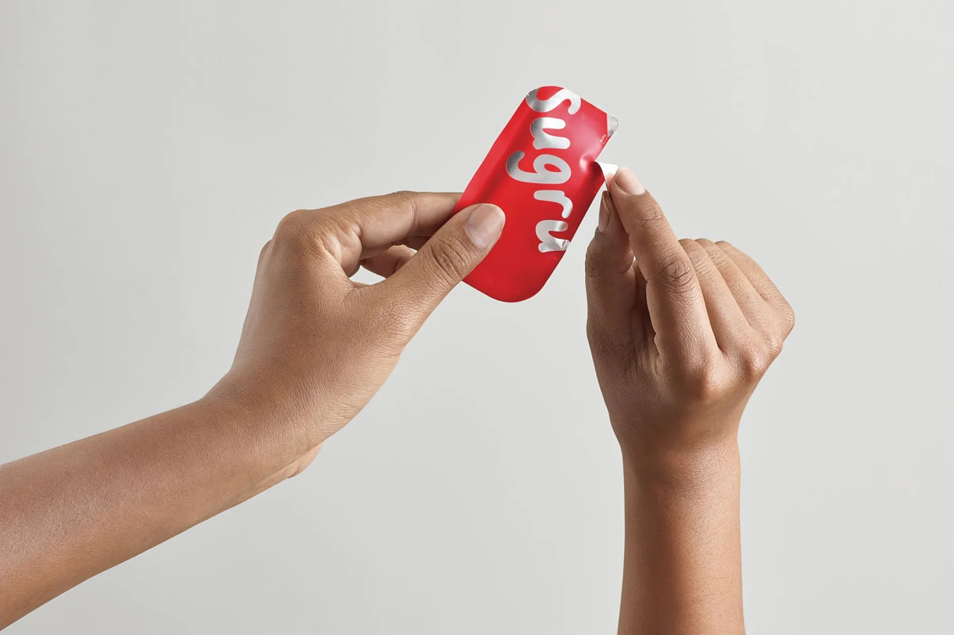 無地の背景に、小さな赤い長方形の Sugru パッケージを両手で持っています。片方の手は、テサ テープを使ってパッケージを破り開け、中身の一部を露出させています。. (This text has been generated by AI)