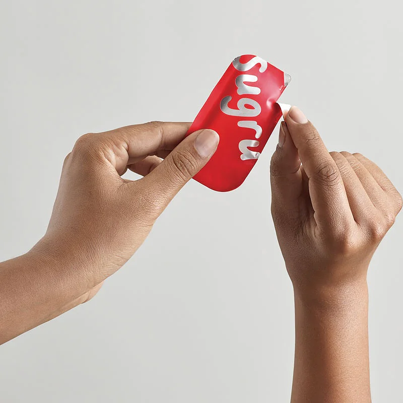 無地の背景に、小さな赤い長方形の Sugru パッケージを両手で持っています。片方の手は、テサ テープを使ってパッケージを破り開け、中身の一部を露出させています。. (This text has been generated by AI)