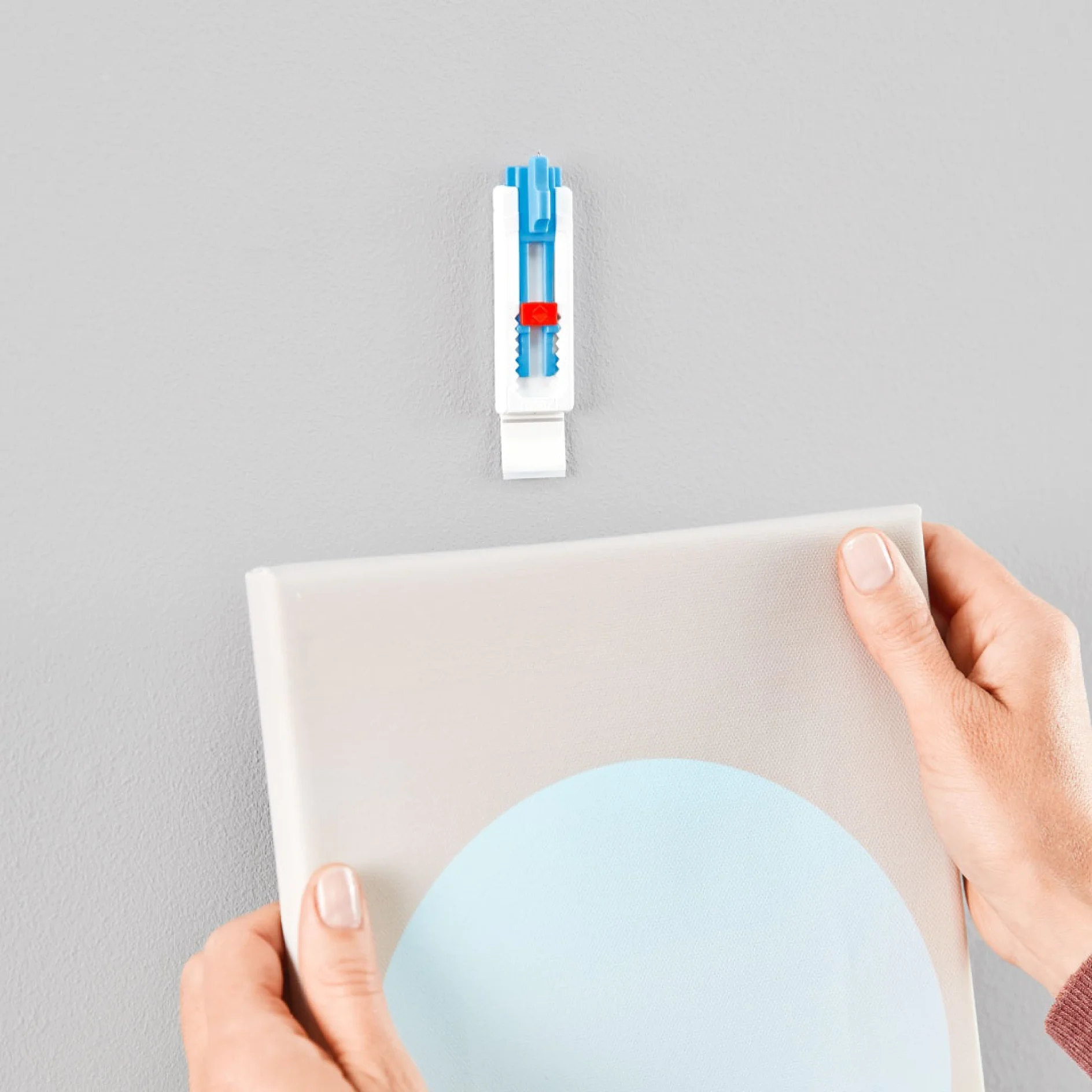 壁の近くに青い円が描かれたキャンバスを持つ手。キャンバスの上には、白と青の壁用ネジが壁に差し込まれており、テサ テープを使用してアートワークを掛ける準備ができています。背景は無地の灰色の壁です。. (This text has been generated by AI)