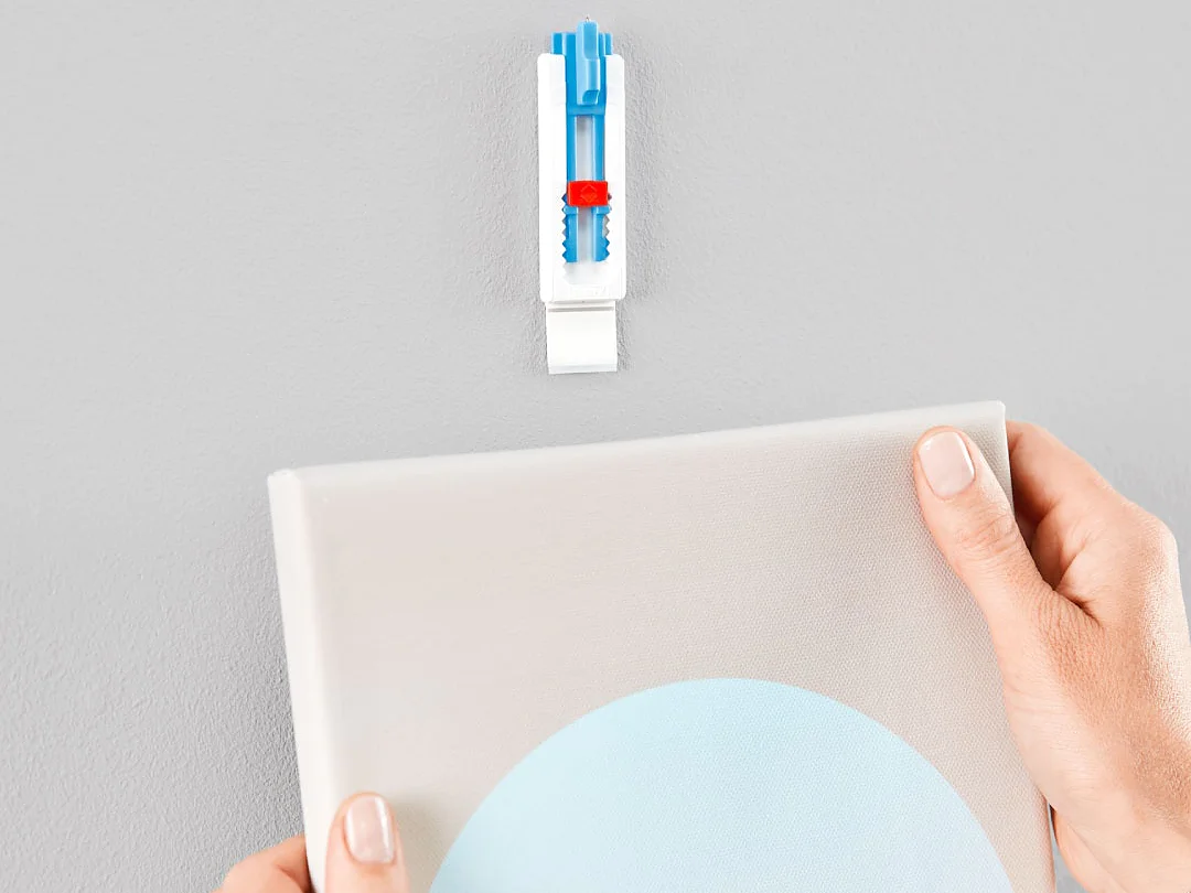 壁の近くに青い円が描かれたキャンバスを持つ手。キャンバスの上には、白と青の壁用ネジが壁に差し込まれており、テサ テープを使用してアートワークを掛ける準備ができています。背景は無地の灰色の壁です。. (This text has been generated by AI)