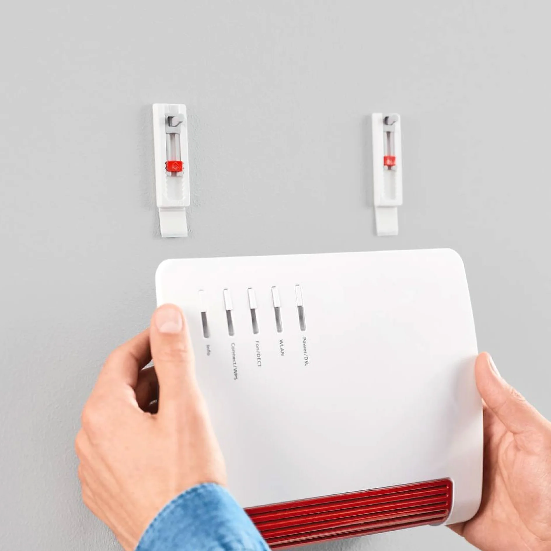 青いシャツを着た人が、テサ テープ クリップを使用して、赤い通気口が付いた白いルーターを壁に取り付けています。ルーターにはインジケータ ライトとボタンが付いています。デバイスをしっかりと固定するために、2 つのクリップが壁に取り付けられています。. (This text has been generated by AI)