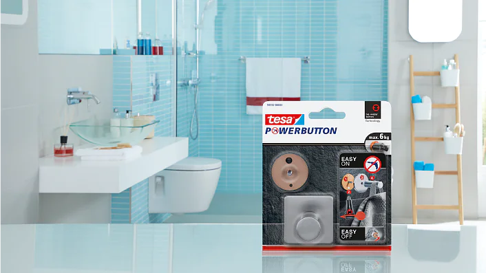 Tesa Powerbutton のパッケージは、モダンなシンク、トイレ、タオルや洗面用具が置かれたはしご型の棚を備えたバスルームのカウンタートップの前景に目立つように展示されています。主な色は青と白です。. (This text has been generated by AI)