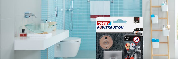 Tesa Powerbutton のパッケージは、モダンなシンク、トイレ、タオルや洗面用具が置かれたはしご型の棚を備えたバスルームのカウンタートップの前景に目立つように展示されています。主な色は青と白です。. (This text has been generated by AI)