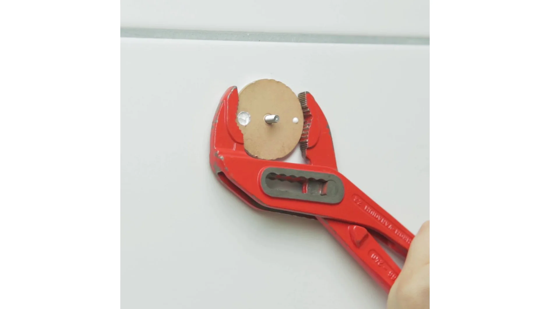 赤いモンキーレンチが、白いタイル張りの壁に、中央にボルトが通っている茶色の円形ディスクを押さえています。レンチを握っている人の手が部分的に見えます。. (This text has been generated by AI)