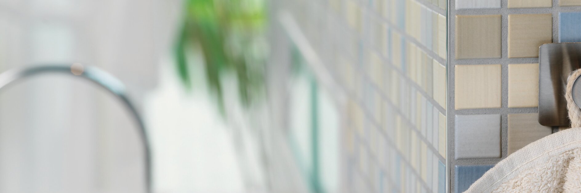 ベージュのタオルが、テサテープを使用してグレー、ベージュ、ブルーの色合いの小さな正方形のタイルが貼られた壁に取り付けられたモダンな金属製のフックに掛けられています。画像の左側には、浴槽の蛇口がぼやけて見えます。. (This text has been generated by AI)