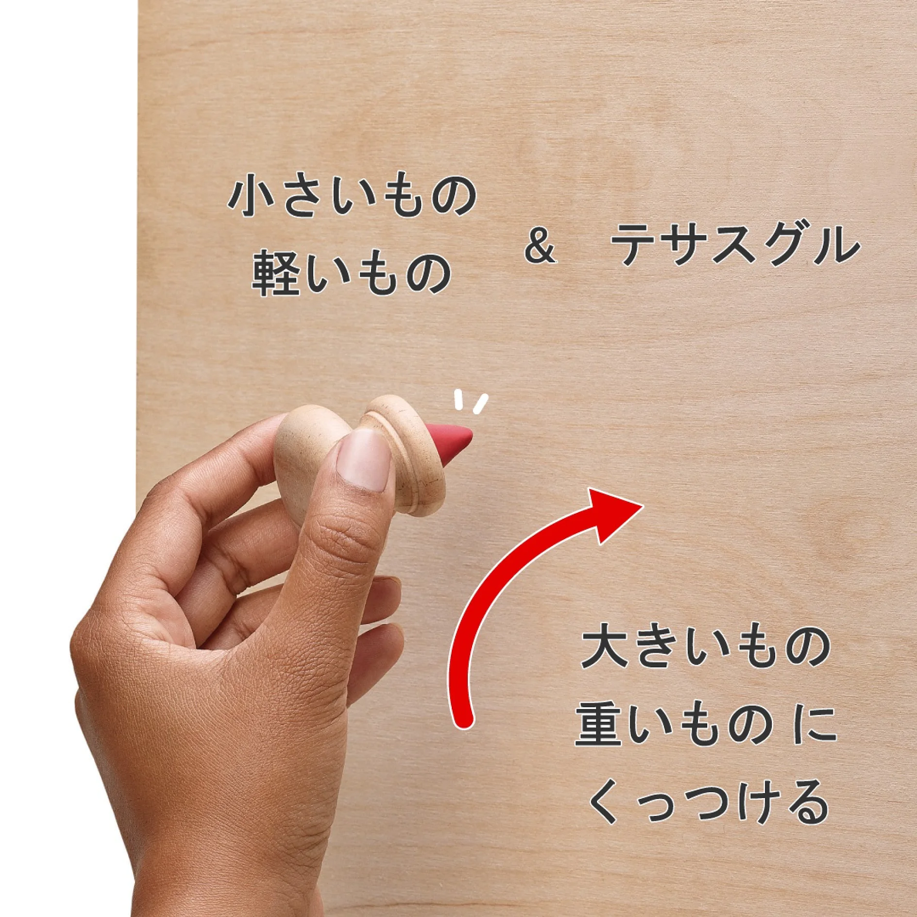 手には、赤い矢印と下向きの日本語の文字が書かれた小さな木製のこまが握られています。背景は明るい木製の表面です。日本語の文字には、テサ テープを使用したオブジェクトに関する説明が記載されています。. (This text has been generated by AI)