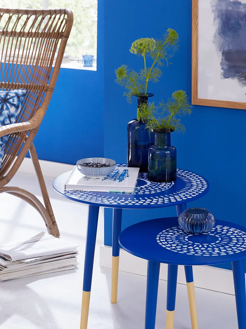 A room featuring a wicker chair with a patterned cushion, two blue nested tables with decorative items, and a vibrant blue wall with a framed abstract painting. A jute rug sits on the white floor, and a stack of books is nearby. (This text has been generated by AI)