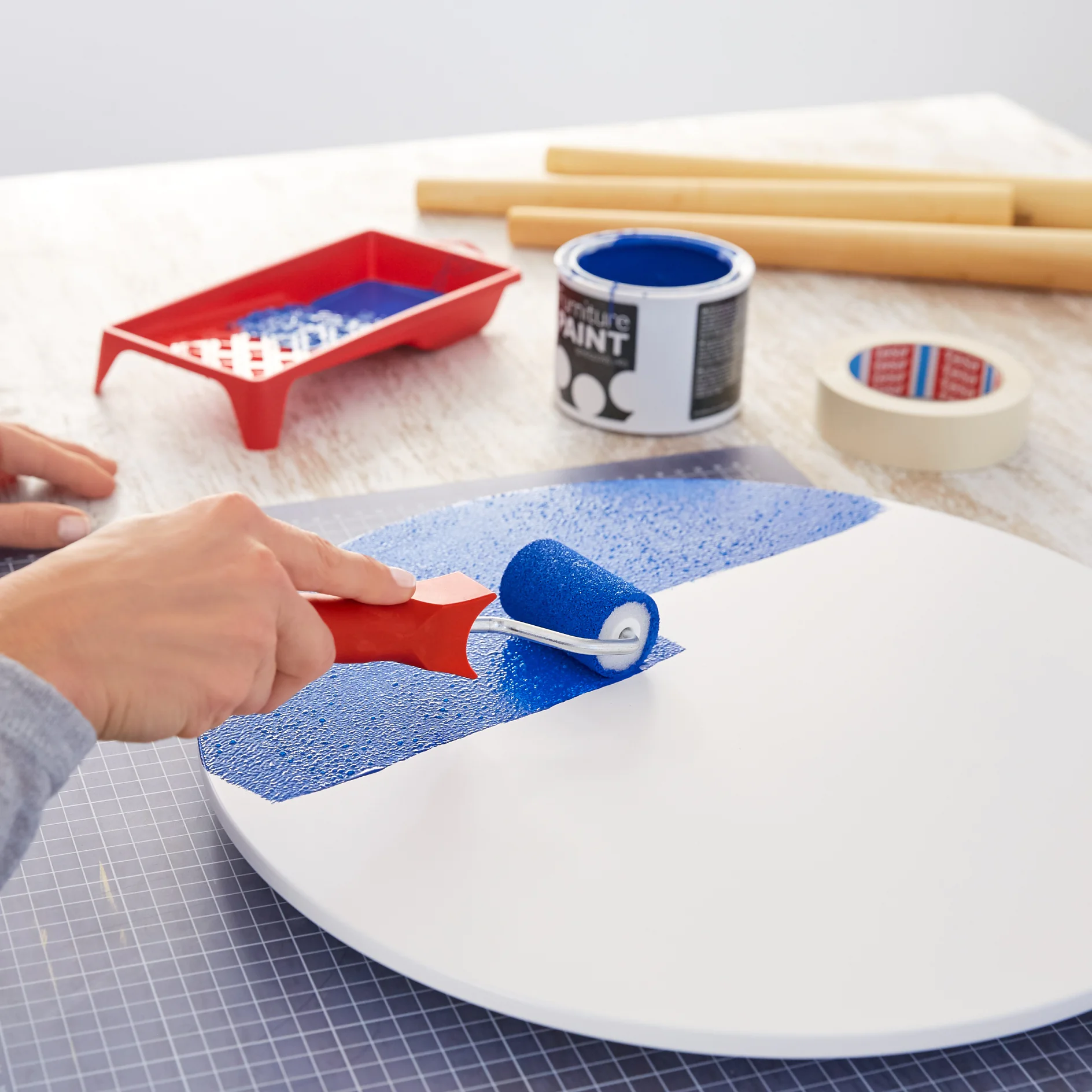 A person uses a small paint roller to apply blue paint onto a white circular surface. A red paint tray, a can of blue paint, tesa tape, and wooden dowels are on the table in the background. (This text has been generated by AI)