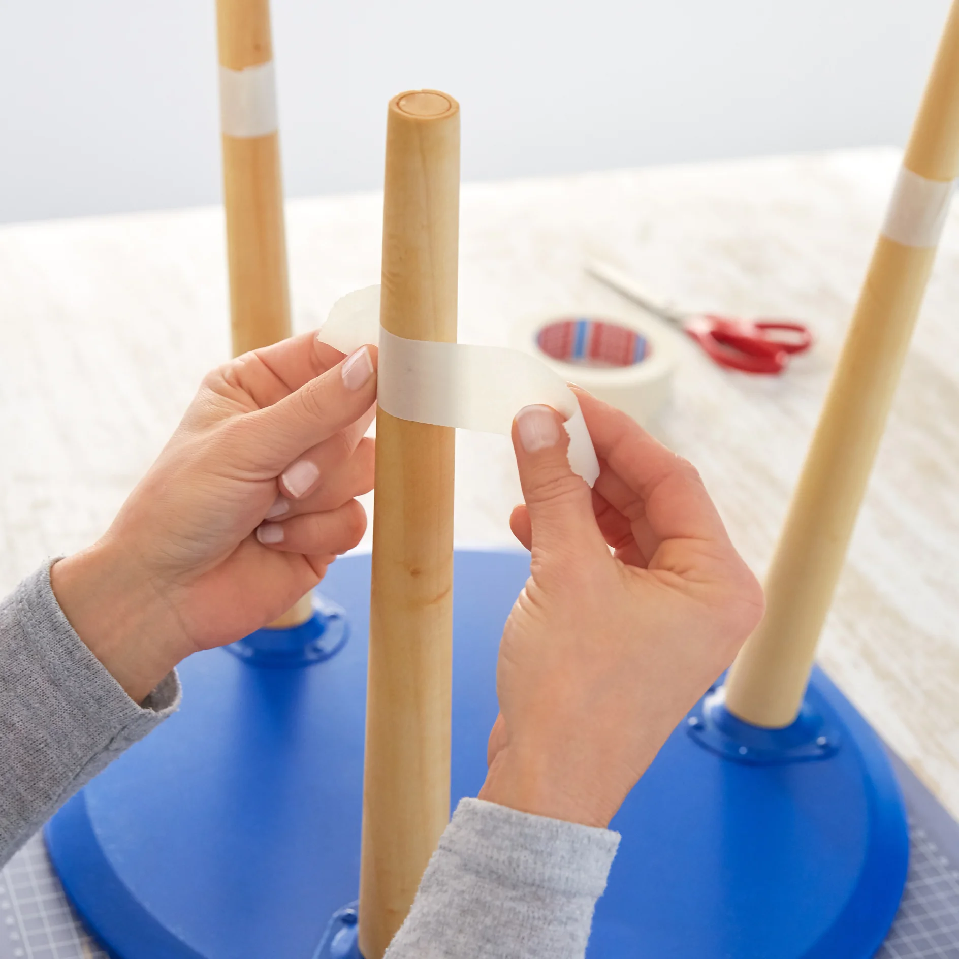 Hands wrapping tesa tape around a wooden table leg attached to an upside-down blue tabletop. In the background, a roll of tesa tape and red scissors are on the light-colored surface. (This text has been generated by AI)