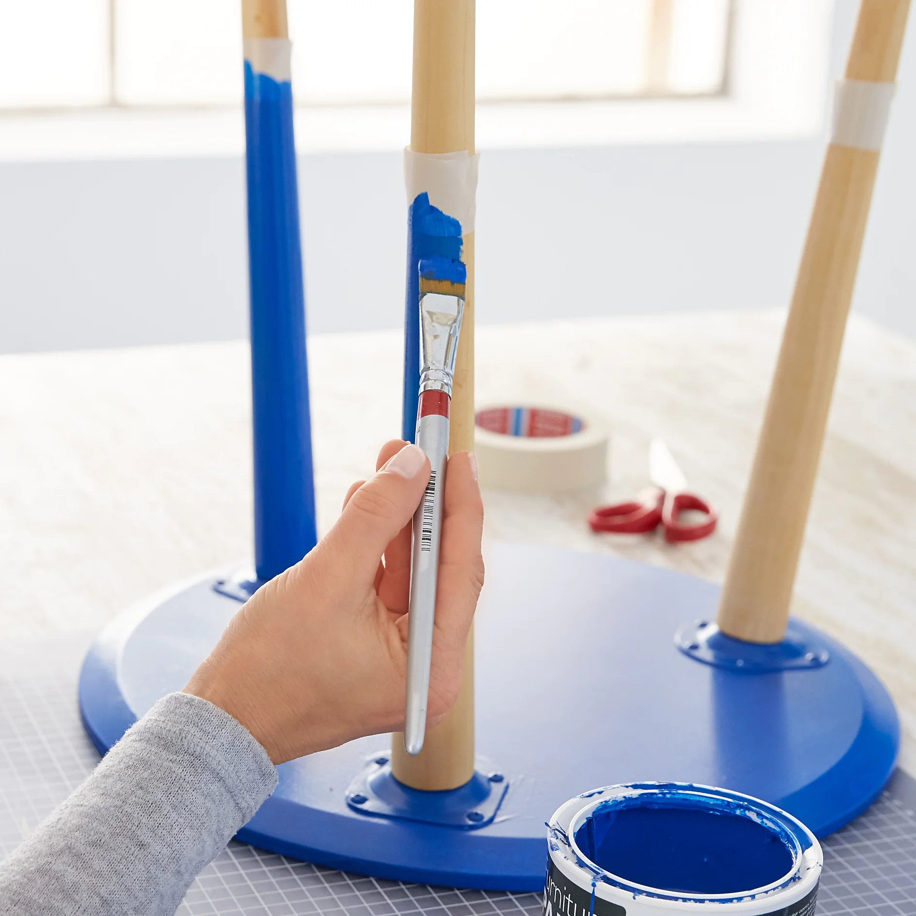 A person is painting a wooden furniture leg blue with a brush. The leg is taped off with tesa tape to create a clean edge. The furniture appears to be upside down on a table. A pair of scissors and tesa masking tape are in the background. (This text has been generated by AI)