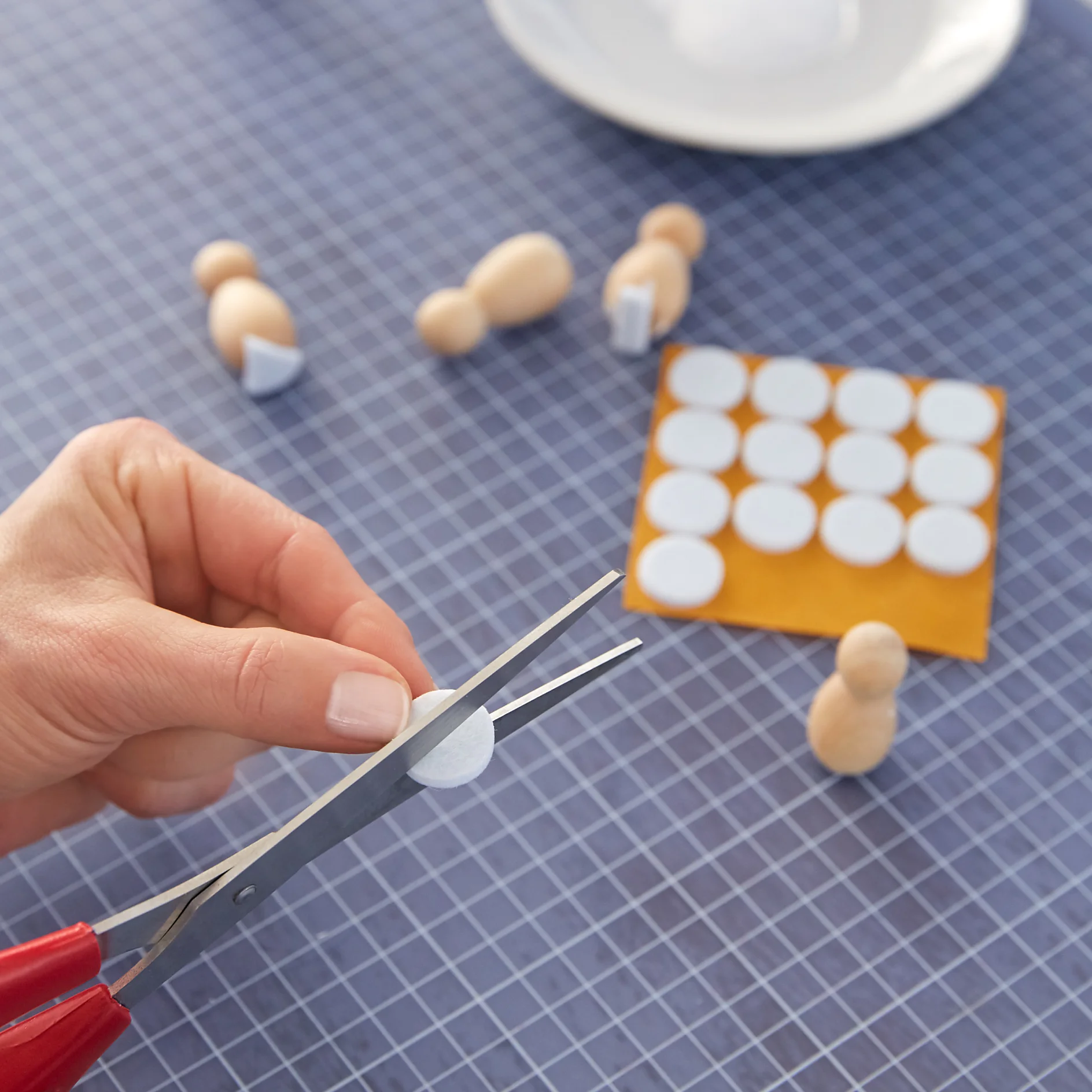 A hand uses scissors to cut a small white felt circle from a larger sheet on a grid-patterned cutting mat. Wooden peg figures, some with half-circles attached using tesa tape, are on the mat. A white plate is partially visible in the background. (This text has been generated by AI)