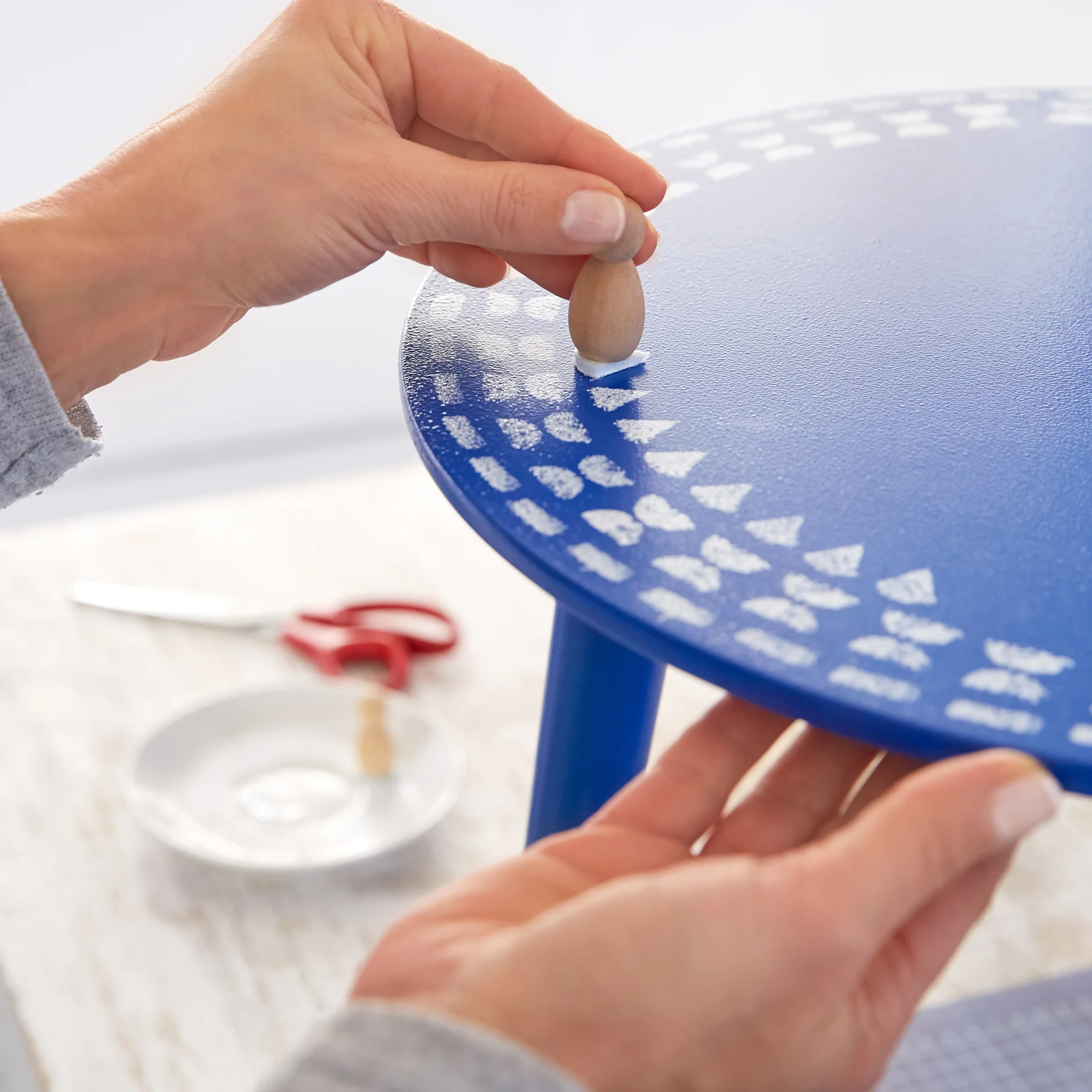A person stamps white triangular patterns onto a blue round table using a small wooden tool. Red scissors, a white plate, and another tesa tape tool rest on a light-colored surface in the background. (This text has been generated by AI)
