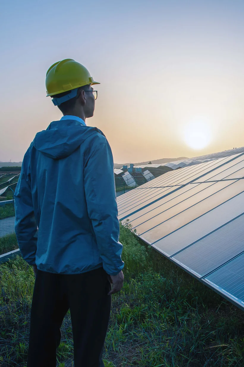 A person in a yellow hard hat and blue jacket stands in a grassy area, looking at a large array of solar panels at sunset. The sky is a gradient of orange and blue, and distant hills are visible under the setting sun. (This text has been generated by AI)