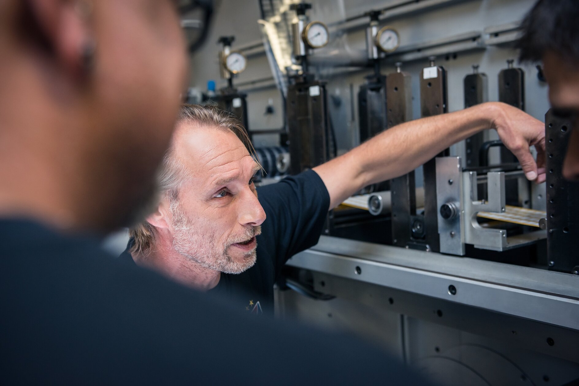 A person gestures towards machinery, explaining something to two others who are partially visible in the foreground. The setting appears to be an industrial or workshop environment with metal equipment and gauges, prominently featuring tesa tape. (This text has been generated by AI)