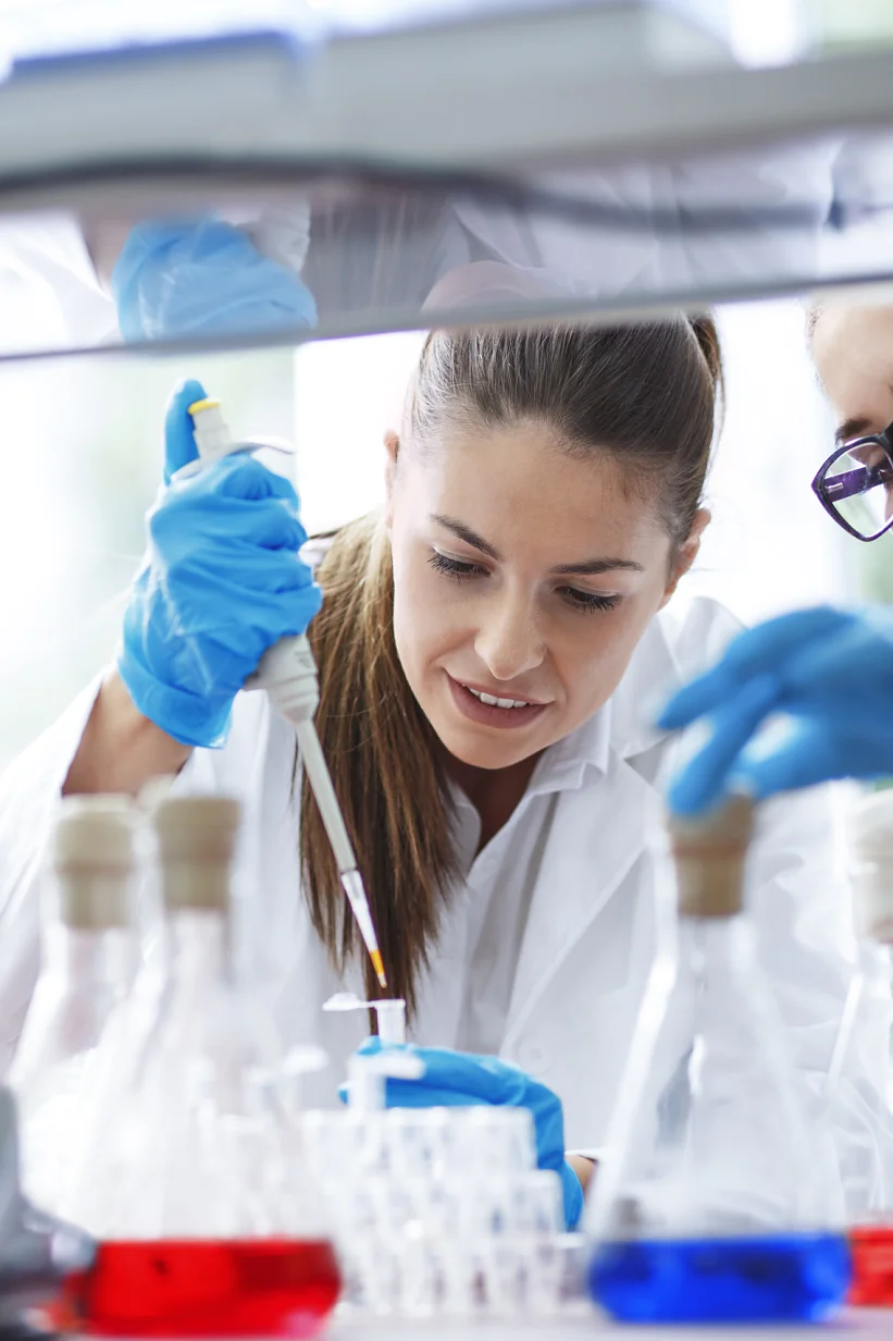 In a laboratory, two scientists wearing lab coats and gloves are hard at work. One carefully uses a pipette while the other watches attentively. The table is filled with various lab equipment and bottles containing colored liquids. Both scientists seem deeply focused and engaged in their experiment, using tesa tape for precision tasks where needed. (This text has been generated by AI)