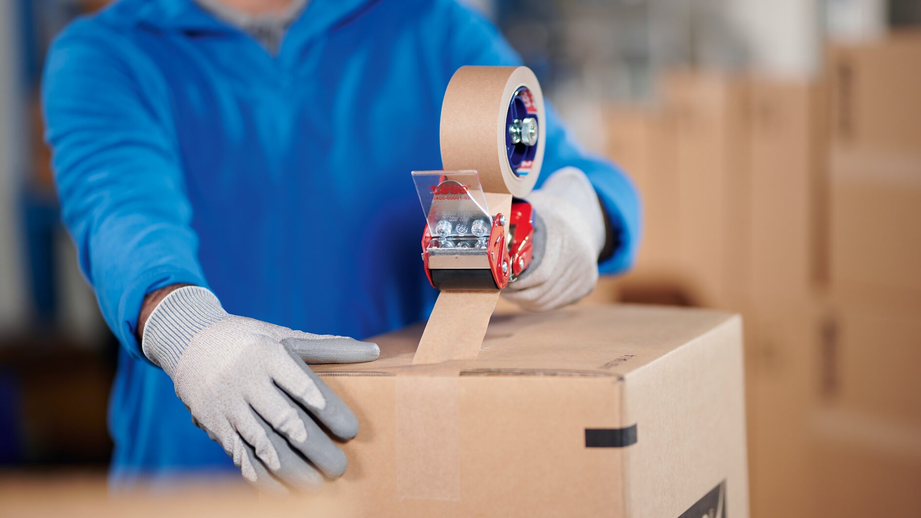A person wearing a blue jacket and gray gloves is sealing a cardboard box with brown packing tesa tape using a red tesa tape dispenser. Other cardboard boxes are visible in the background. (This text has been generated by AI)