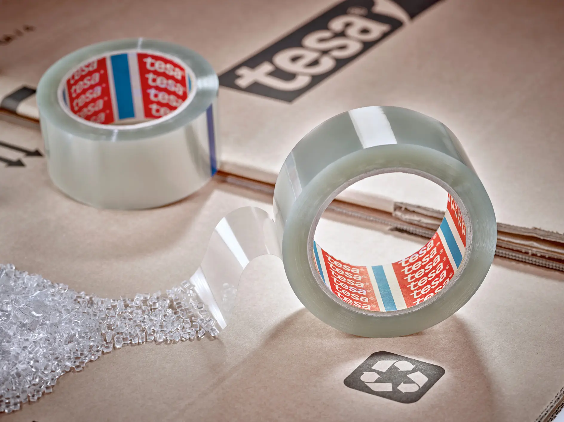 A roll of transparent tesa tape with a red and white inner label is partially unrolled on top of a cardboard box with a recycling symbol. Bubble wrap is nearby. Another roll of tesa tape is in the background. The cardboard has tesa printed on it.