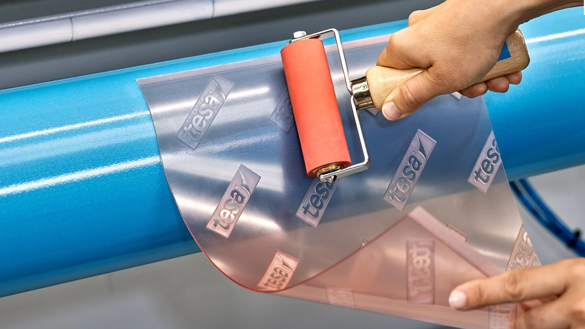 手は赤いゴムローラーを使用して、「tesa テープ」と表示された透明な粘着シートを青い円筒形の表面に貼り付けます。. (This text has been generated by AI)