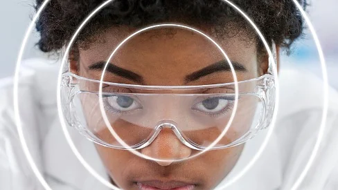A person wearing safety goggles is looking intently at the camera. Concentric white circles are superimposed over the image, centered on their face. The person has short curly hair and is wearing a white garment. In their hand, they hold a roll of tesa tape, ready for use in securing materials or during a home project. (This text has been generated by AI)