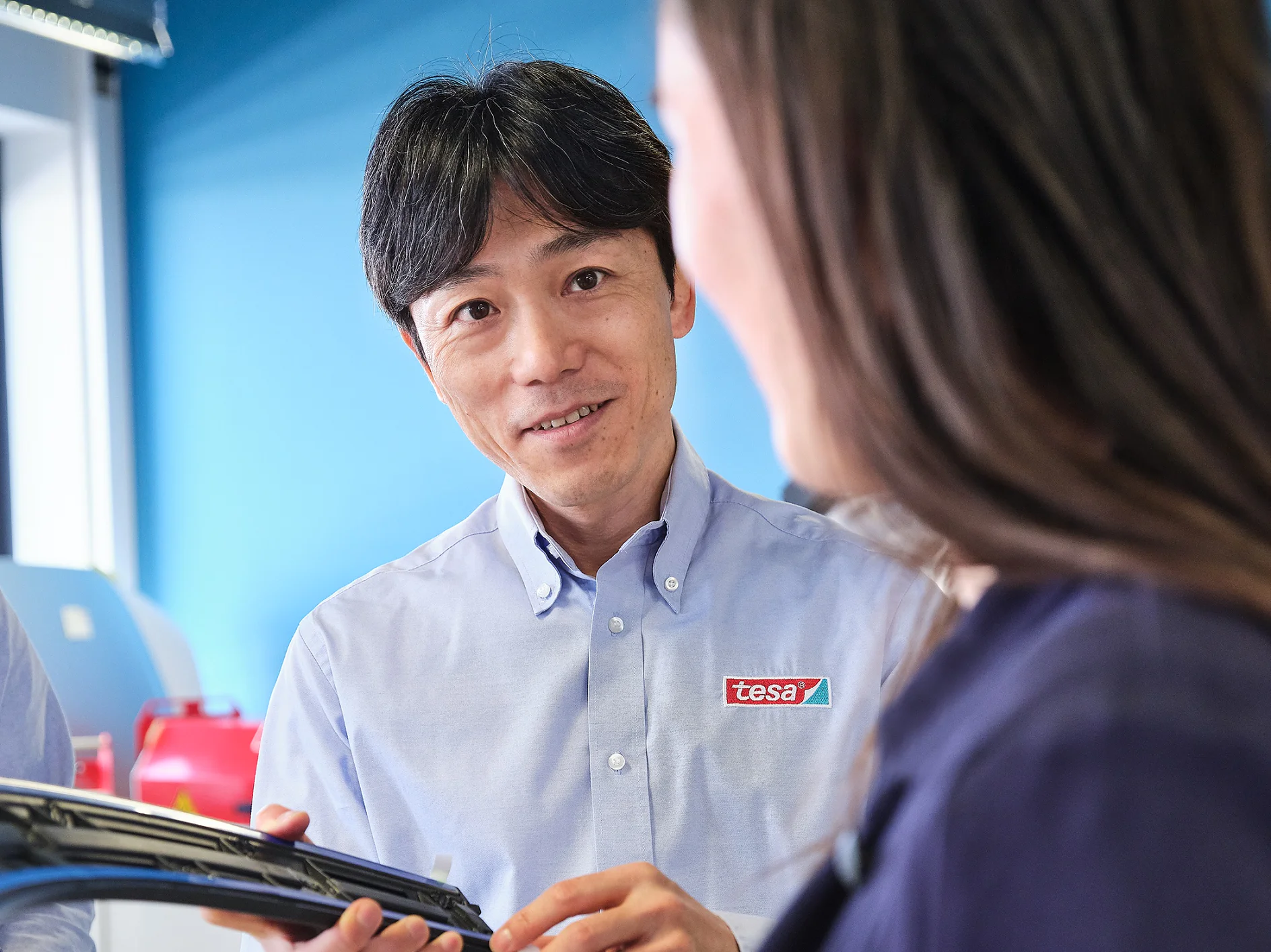 A man in a blue shirt with a tesa logo holds an object, speaking to a woman with long hair. They are in a brightly lit room with a blue wall. (This text has been generated by AI)