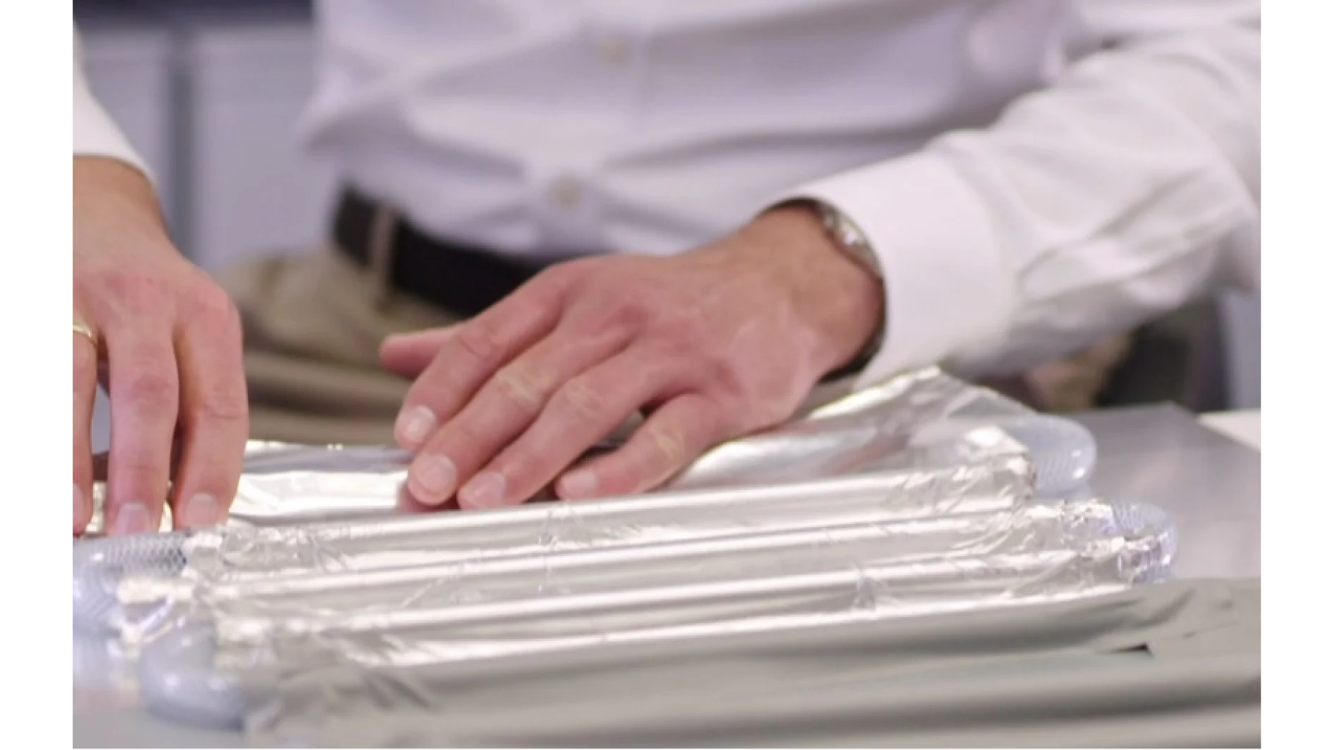 A person in a white shirt is arranging multiple shiny, cylindrical objects wrapped in aluminum foil on a table. The persons hands are visible, gently handling the objects secured with tesa tape. The background is blurred. (This text has been generated by AI)